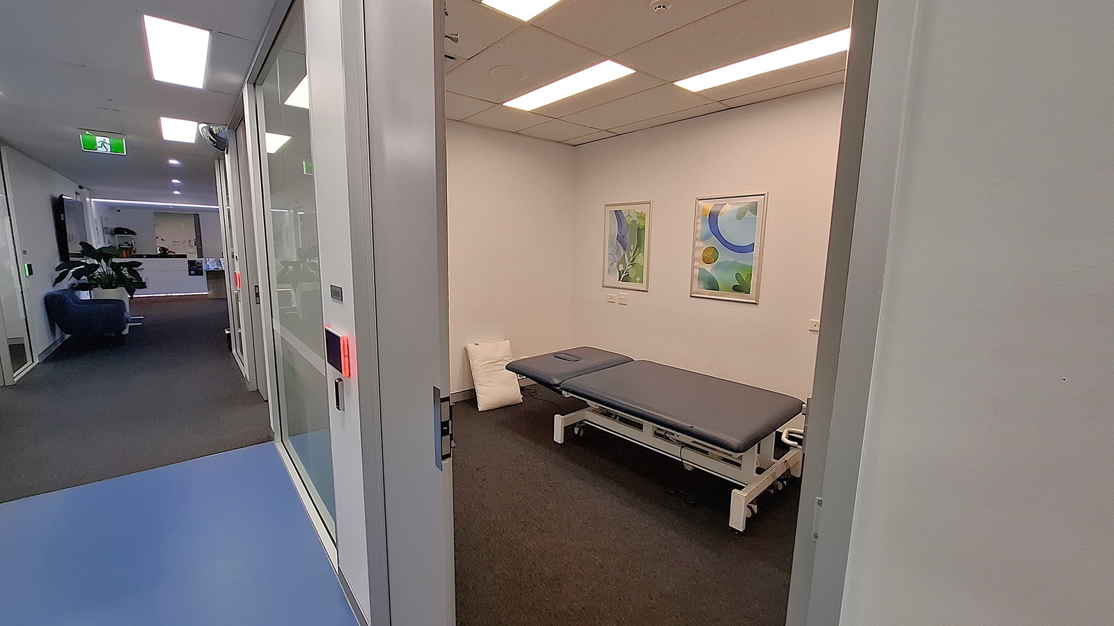 On the left, a hallway with a blue floor and glass walls, leading to a reception area with a blue sofa and potted plants. In the middle, an open door reveals a room with a grey examination table, two framed abstract artworks on the wall, and a white pillow on the floor. On the right, a white wall with part of the door frame visible. The floor in the room is covered with dark carpet, and the ceiling has white panels with recessed lighting. On the left, a hallway with a blue floor and glass walls, leading to a reception area with a blue sofa and potted plants. In the middle, an open door reveals a room with a grey examination table, two framed abstract artworks on the wall, and a white pillow on the floor. On the right, a white wall with part of the door frame visible. The floor in the room is covered with dark carpet, and the ceiling has white panels with recessed lighting.