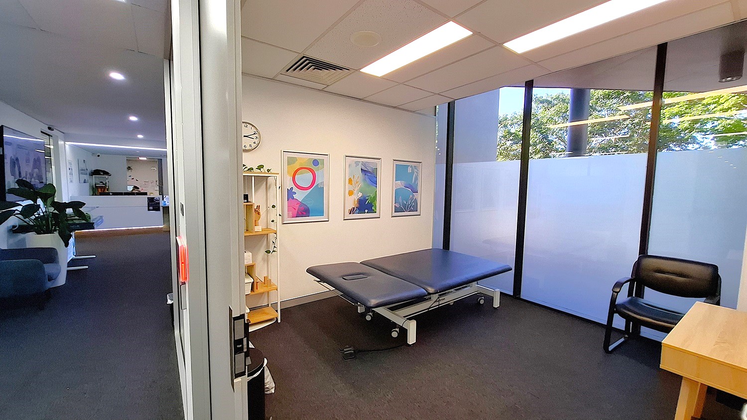 On the left, a hallway with dark carpet, a blue sofa, and potted plants. In the middle, a room with a black examination table, three framed abstract artworks on the wall, a white clock, and a white shelving unit with various items. On the right, large windows with frosted lower panels, a black chair, and a wooden desk. The ceiling has white panels with recessed lighting and air vents. On the left, a hallway with dark carpet, a blue sofa, and potted plants. In the middle, a room with a black examination table, three framed abstract artworks on the wall, a white clock, and a white shelving unit with various items. On the right, large windows with frosted lower panels, a black chair, and a wooden desk. The ceiling has white panels with recessed lighting and air vents.