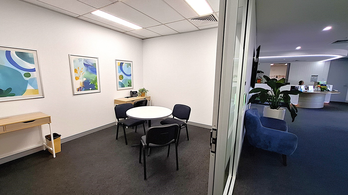 On the left, a small wooden table with a drawer and a bin underneath, three framed abstract artworks on the wall, and a round white table with four black chairs. In the middle, a glass door leading to a hallway. On the right, a blue sofa and a large potted plant. In the background, a curved reception desk with a person seated behind it and informational brochures. The floor is covered with dark carpet, and the ceiling has white panels with recessed lighting and air vents. On the left, a small wooden table with a drawer and a bin underneath, three framed abstract artworks on the wall, and a round white table with four black chairs. In the middle, a glass door leading to a hallway. On the right, a blue sofa and a large potted plant. In the background, a curved reception desk with a person seated behind it and informational brochures. The floor is covered with dark carpet, and the ceiling has white panels with recessed lighting and air vents.