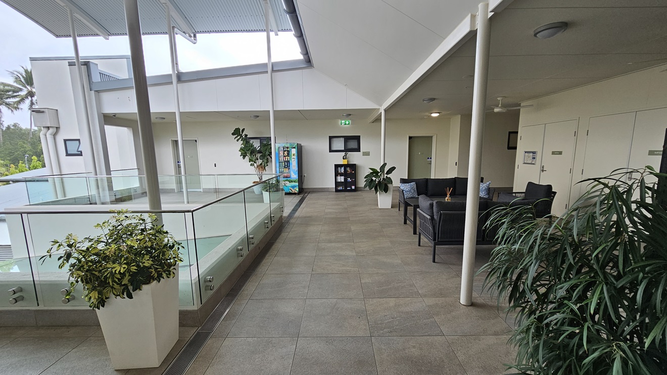 To the right is a seating area with black cushioned chairs, adorned with plants. Glass railings secure the space, leading to a vending machine offering cold drinks at the far end. Next to it is a wooden cabinet with books. A raised corrugated roof provides additional shade. The walls are white, and the floor has grey tiles. To the right is a seating area with black cushioned chairs, adorned with plants. Glass railings secure the space, leading to a vending machine offering cold drinks at the far end. Next to it is a wooden cabinet with books. A raised corrugated roof provides additional shade. The walls are white, and the floor has grey tiles.