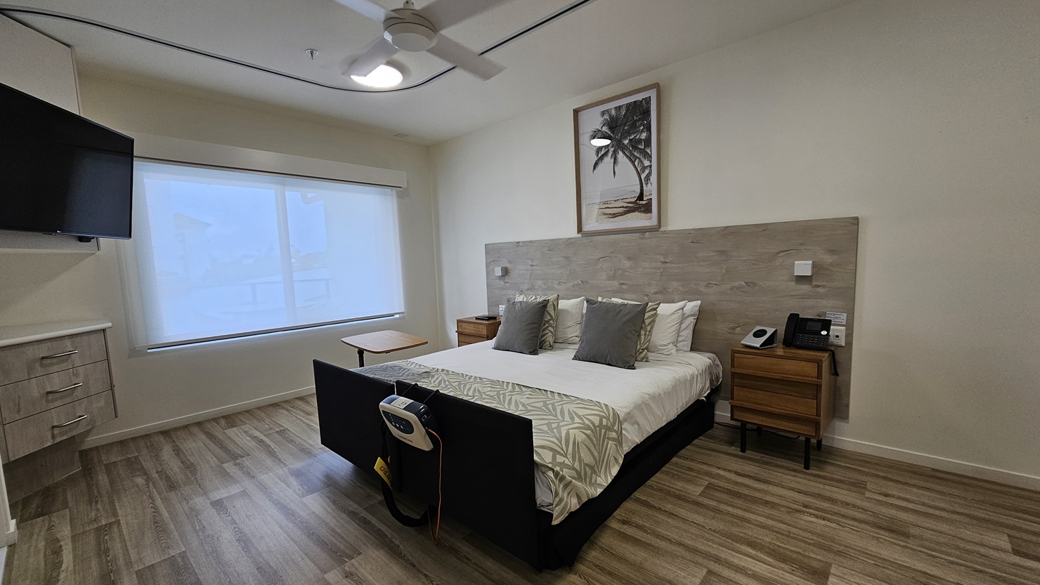 In the middle is a double bed with a wooden headrest. Wooden cabinets flank both sides, one holding a telephone. A painting hangs above the headrest. To the left is a large window with a flatscreen TV mounted on the wall. Below it is another wooden cabinet. The floor is timber, and the wall and ceiling are white with an LED light and a ceiling fan. In the middle is a double bed with a wooden headrest. Wooden cabinets flank both sides, one holding a telephone. A painting hangs above the headrest. To the left is a large window with a flatscreen TV mounted on the wall. Below it is another wooden cabinet. The floor is timber, and the wall and ceiling are white with an LED light and a ceiling fan.
