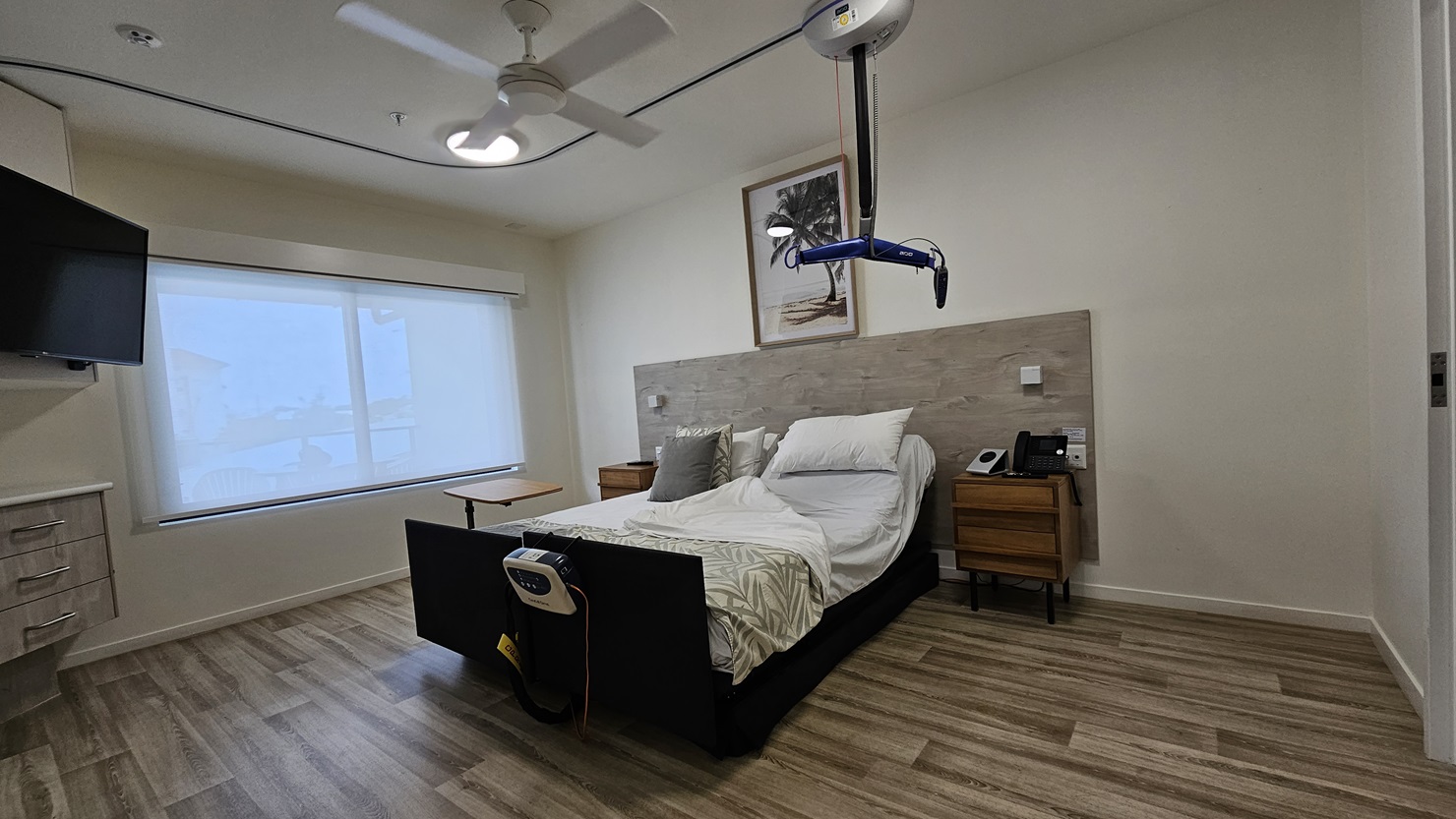 In the middle is a double bed with a wooden bedhead. Wooden cabinets flank both sides, one holding a telephone. A painting hangs above the headrest. A hoist hangs from the ceiling, and a monitor is attached to the bed. To the left is a large window with a flatscreen TV mounted on the wall. Below it is another wooden cabinet. The floor is timber, and the wall and ceiling are white with an LED light and a ceiling fan. In the middle is a double bed with a wooden bedhead. Wooden cabinets flank both sides, one holding a telephone. A painting hangs above the headrest. A hoist hangs from the ceiling, and a monitor is attached to the bed. To the left is a large window with a flatscreen TV mounted on the wall. Below it is another wooden cabinet. The floor is timber, and the wall and ceiling are white with an LED light and a ceiling fan.