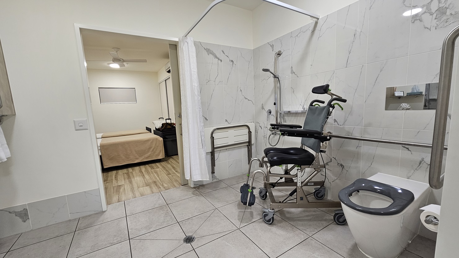 In the ensuite bathroom, an aquatic wheelchair sits in the corner near the shower. A foldable bench chair is mounted to the wall, with a curtain hanging from above. To the right is a toilet bowl equipped with handrails and a tissue dispenser, with a mirror above it. The walls have white tiles, and the ceiling is also white. On the left is the bedroom area. In the ensuite bathroom, an aquatic wheelchair sits in the corner near the shower. A foldable bench chair is mounted to the wall, with a curtain hanging from above. To the right is a toilet bowl equipped with handrails and a tissue dispenser, with a mirror above it. The walls have white tiles, and the ceiling is also white. On the left is the bedroom area.