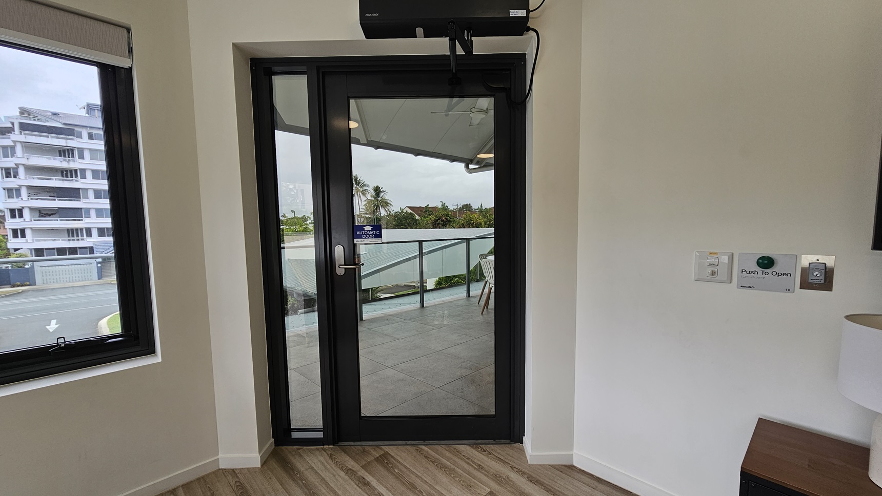 Access the accommodation through an automatic glass door with a "Push To Open" button on the right. A TV stand and a white lampshade stand on the right. To the left is a window offering a view of the outside. The floor is made of timber, and the walls are all white. Access the accommodation through an automatic glass door with a "Push To Open" button on the right. A TV stand and a white lampshade stand on the right. To the left is a window offering a view of the outside. The floor is made of timber, and the walls are all white.