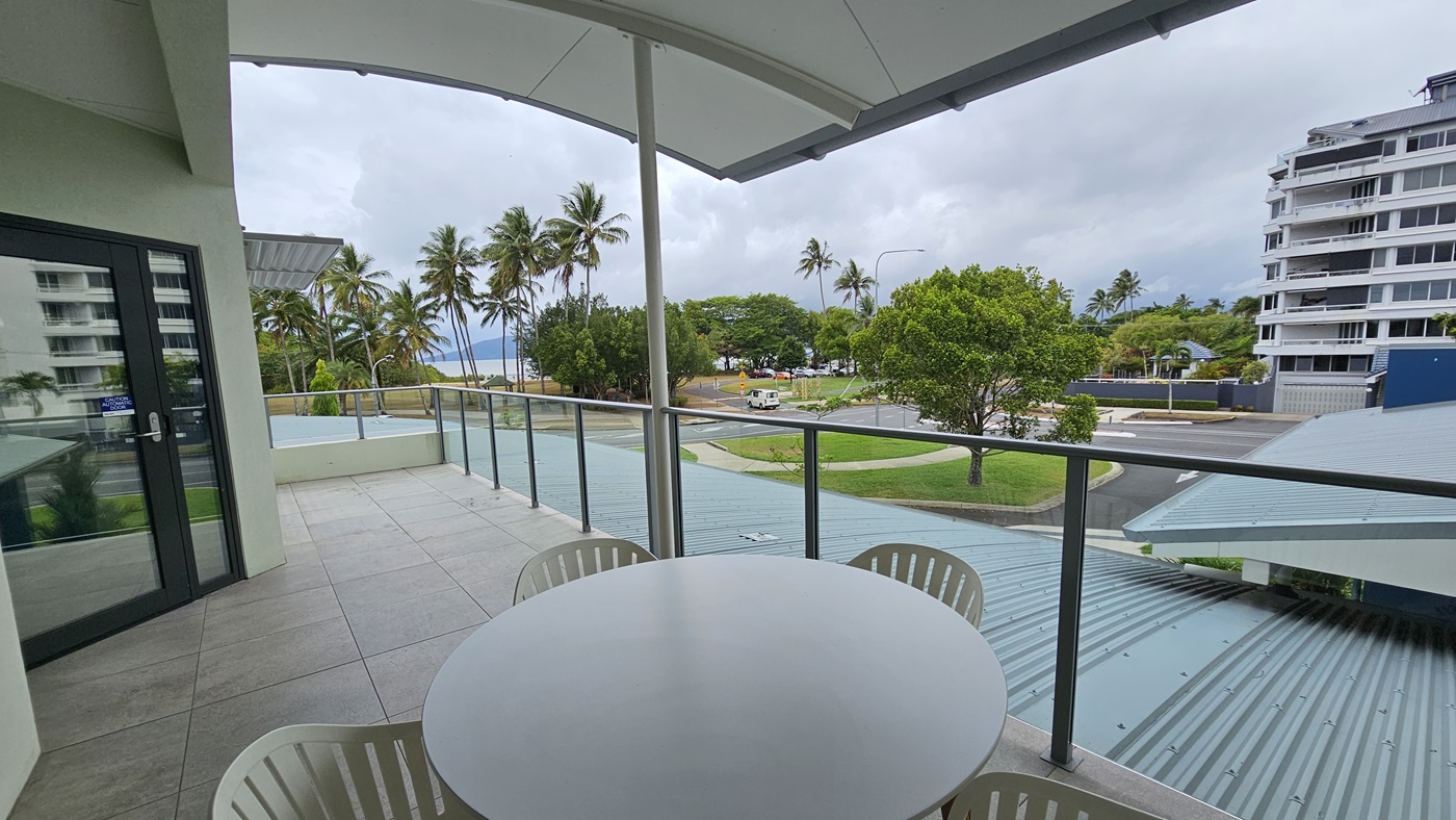 The balcony features a round table and four movable plastic chairs in the middle. The roof extends for additional shade, with glass railings securing the area. To the left is a glass door, while tall palm trees line the left side along with other trees. To the right is the road and a large building. The balcony features a round table and four movable plastic chairs in the middle. The roof extends for additional shade, with glass railings securing the area. To the left is a glass door, while tall palm trees line the left side along with other trees. To the right is the road and a large building.