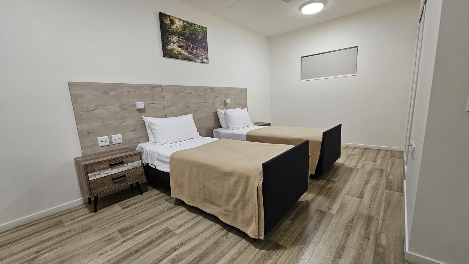In the middle are two single beds placed side by side, each with a bedside table and a wooden headrest. A painting hangs above. The walls and ceilings are all white with LED lights. The floor is made of timber. In the middle are two single beds placed side by side, each with a bedside table and a wooden headrest. A painting hangs above. The walls and ceilings are all white with LED lights. The floor is made of timber.