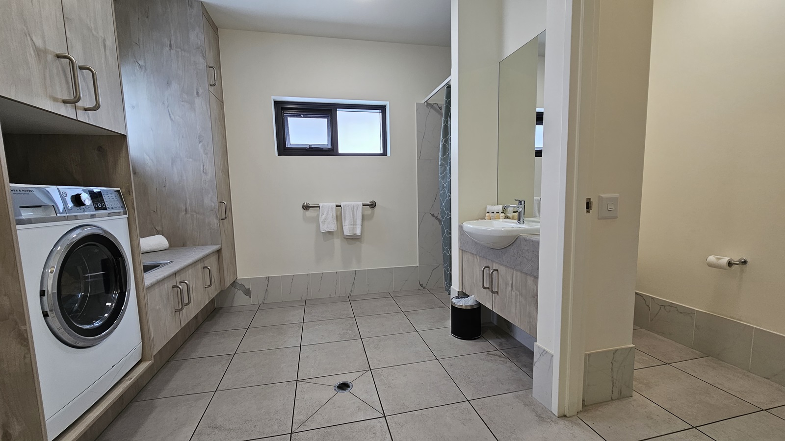The laundry area has a washing machine on the left and a sink beside it. Two small windows are in the middle, with a hand grab and two white towels below. On the right is a wash basin, toilet bowl, and shower area. A rubbish bin is placed under the wash basin. The floor has grey tiles, and the ceiling and walls are all white. The laundry area has a washing machine on the left and a sink beside it. Two small windows are in the middle, with a hand grab and two white towels below. On the right is a wash basin, toilet bowl, and shower area. A rubbish bin is placed under the wash basin. The floor has grey tiles, and the ceiling and walls are all white.