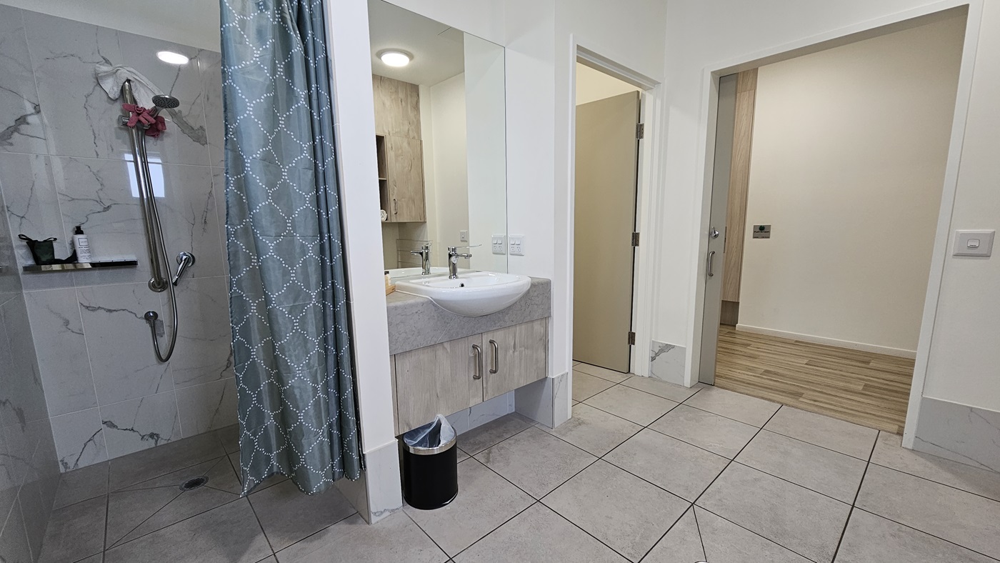 On the right is a shower with a green shower curtain and a shampoo holder. Beside it is a washbasin with a large mirror. Underneath is a black rubbish bin. Adjacent is an inward-opening door. The floor has grey tiles, and the ceiling and walls are all white with a black accent. To the right is a door and a timber floor is visible. On the right is a shower with a green shower curtain and a shampoo holder. Beside it is a washbasin with a large mirror. Underneath is a black rubbish bin. Adjacent is an inward-opening door. The floor has grey tiles, and the ceiling and walls are all white with a black accent. To the right is a door and a timber floor is visible.