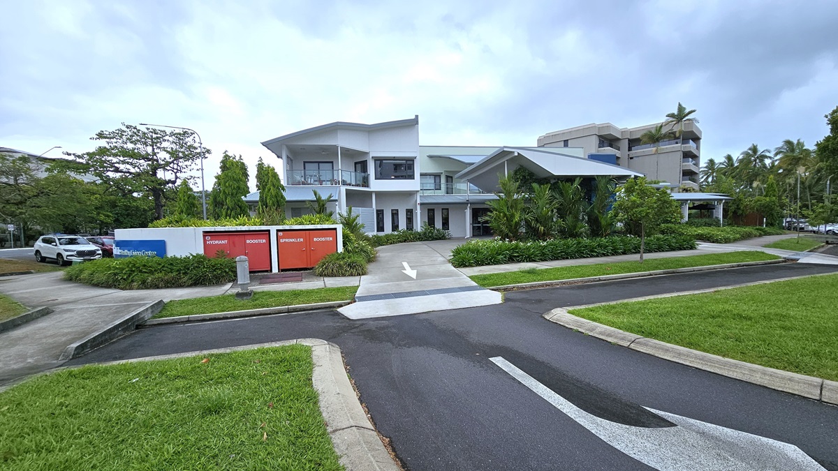On the left are parked vehicles on the side of the road. A concrete pathway extends from the car park to the entrance. In the centre stands a white concrete fence displaying the facility logo "Spinal Life Healthy Living Cairns". Behind it, a modern two-story building crafted from glass and concrete is nestled amidst trees, landscaped ornamental plants, and green grass. Step-free ramps are accessible. On the left are parked vehicles on the side of the road. A concrete pathway extends from the car park to the entrance. In the centre stands a white concrete fence displaying the facility logo "Spinal Life Healthy Living Cairns". Behind it, a modern two-story building crafted from glass and concrete is nestled amidst trees, landscaped ornamental plants, and green grass. Step-free ramps are accessible.