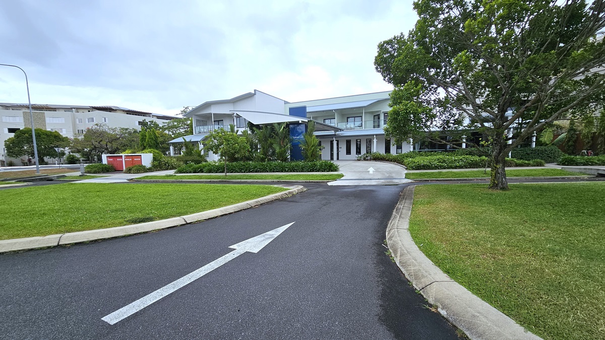 A bitumen road, marked with white arrows, directs towards a concrete pathway to a two-story white building. Ahead, landscaped ornamental plants, manicured grass, and various trees adorn the surroundings. Manicured grass stretches towards the front near the parking area. To the far left, a large white building and tall street lights are visible. A bitumen road, marked with white arrows, directs towards a concrete pathway to a two-story white building. Ahead, landscaped ornamental plants, manicured grass, and various trees adorn the surroundings. Manicured grass stretches towards the front near the parking area. To the far left, a large white building and tall street lights are visible.