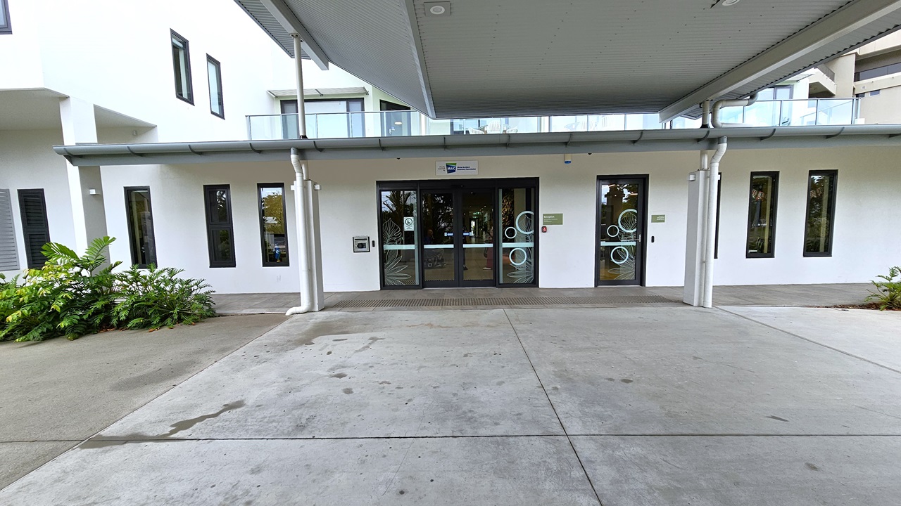 Approaching the building's entrance, a corrugated roof offers shade over the concourse. Ornamental plants adorn the front. Truncated domes mark the floor before the automatic glass sliding door. The building is painted white, with rectangular glass windows. A terrace with a glass railing from the rooftop is also visible. Approaching the building's entrance, a corrugated roof offers shade over the concourse. Ornamental plants adorn the front. Truncated domes mark the floor before the automatic glass sliding door. The building is painted white, with rectangular glass windows. A terrace with a glass railing from the rooftop is also visible.