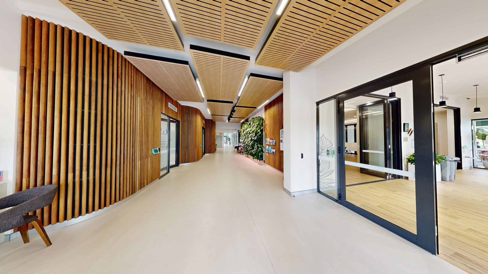 To the right is a sliding door adorned with white floral decoration, leading to the meeting room with timber flooring. The lobby offers access to accessible toilets and other facilities. Timber slats adorn the walls and ceiling, with fluorescent lights providing illumination. Brochures are displayed on a rack near robust indoor plants. The floor is covered in firm white vinyl. To the right is a sliding door adorned with white floral decoration, leading to the meeting room with timber flooring. The lobby offers access to accessible toilets and other facilities. Timber slats adorn the walls and ceiling, with fluorescent lights providing illumination. Brochures are displayed on a rack near robust indoor plants. The floor is covered in firm white vinyl.