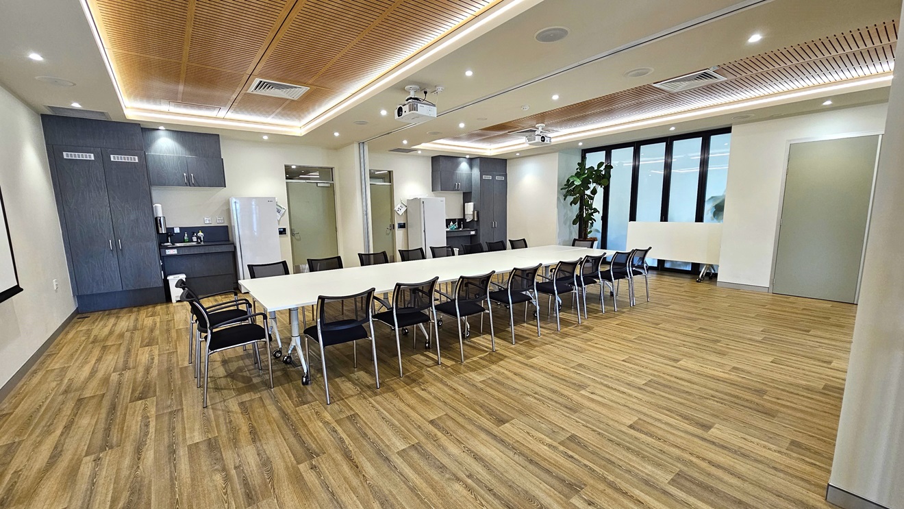 The meeting room has a long white table with black movable chairs. A projector hangs on the left wall. In the middle, a dark grey wooden cabinet, fridge, and wash area are positioned. To the right is a glass wall, accompanied by an indoor plant. The floor is made of timber, and the ceiling is white with wooden slat accents. A projector is mounted on the ceiling The meeting room has a long white table with black movable chairs. A projector hangs on the left wall. In the middle, a dark grey wooden cabinet, fridge, and wash area are positioned. To the right is a glass wall, accompanied by an indoor plant. The floor is made of timber, and the ceiling is white with wooden slat accents. A projector is mounted on the ceiling