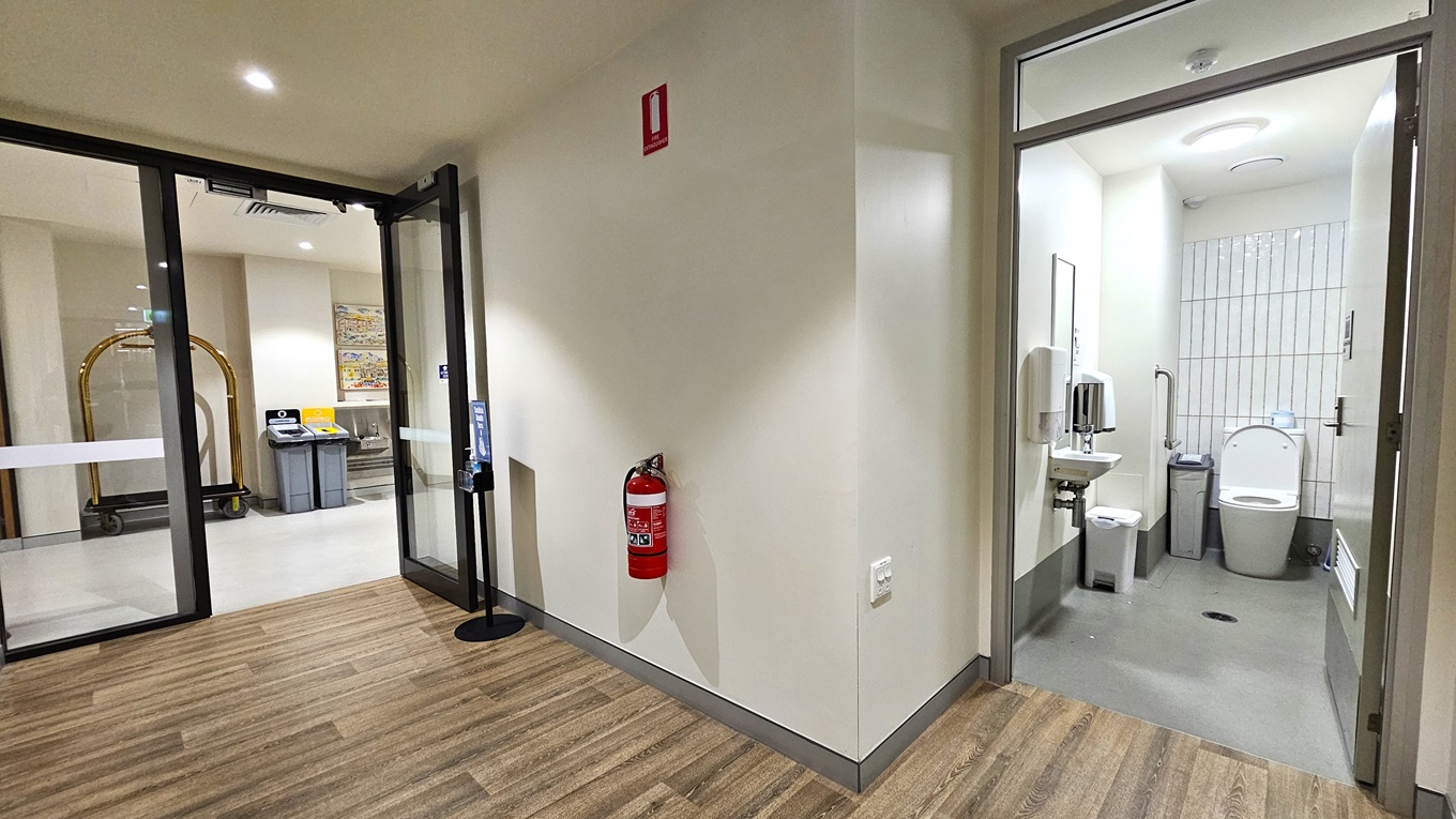 Beside two rubbish bins stands a golden luggage trolley. An inward-opening glass door provides access to accessible toilets, with hand sanitizer and a fire extinguisher nearby. Outside, the floor is timber, changing to grey tiles inside the toilet. Inside, a toilet bowl is in the corner, with rubbish bins nearby. On the left wall, a wash basin, soap, and tissue dispenser are mounted. The ceiling and walls are white. Beside two rubbish bins stands a golden luggage trolley. An inward-opening glass door provides access to accessible toilets, with hand sanitizer and a fire extinguisher nearby. Outside, the floor is timber, changing to grey tiles inside the toilet. Inside, a toilet bowl is in the corner, with rubbish bins nearby. On the left wall, a wash basin, soap, and tissue dispenser are mounted. The ceiling and walls are white.