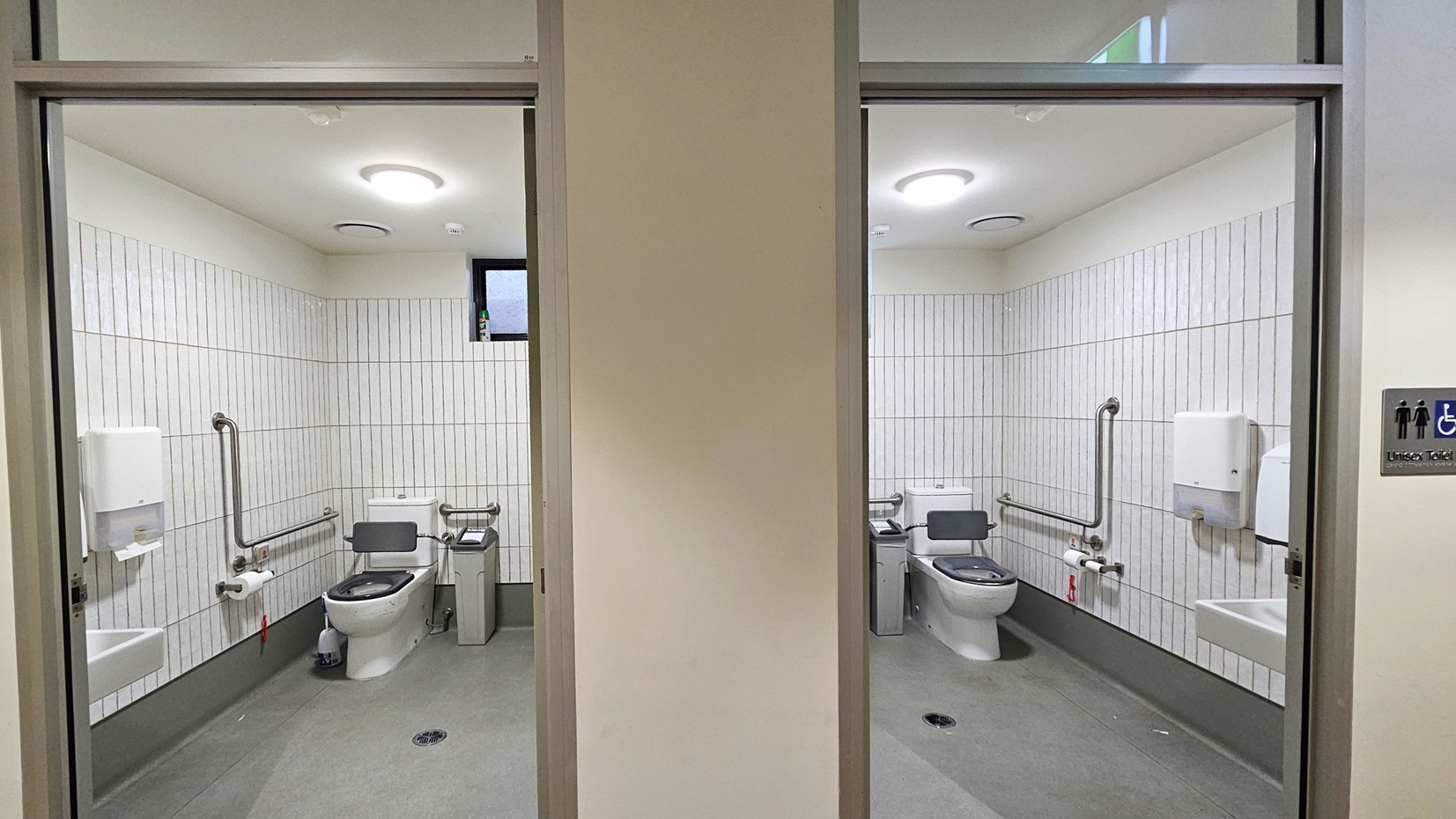 The two toilets feature identical accessories. A toilet bowl is positioned in the corner, alongside a rubbish bin, hand grabs, and a tissue dispenser. A wash basin is mounted on the wall, with a tissue dispenser next to it. The walls have white tiles, and the ceiling is white with LED lights. The two toilets feature identical accessories. A toilet bowl is positioned in the corner, alongside a rubbish bin, hand grabs, and a tissue dispenser. A wash basin is mounted on the wall, with a tissue dispenser next to it. The walls have white tiles, and the ceiling is white with LED lights.