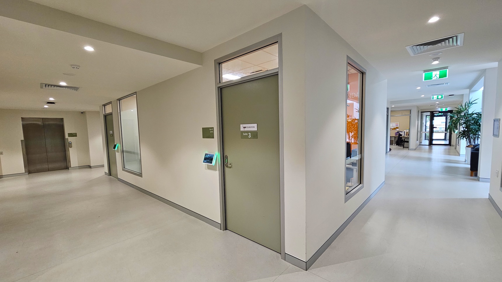 The lobby turns left, leading to two green doors marked with Suite #3 and 4. A lift is on the left. To the right, it leads to a lounge area, Suite #6, and accessible toilets. An indoor plant decorates the lounge. Exit signs hang from the ceiling, continuing towards the glass door. The walls and ceiling are all white and the floor has white vinyl. The lobby turns left, leading to two green doors marked with Suite #3 and 4. A lift is on the left. To the right, it leads to a lounge area, Suite #6, and accessible toilets. An indoor plant decorates the lounge. Exit signs hang from the ceiling, continuing towards the glass door. The walls and ceiling are all white and the floor has white vinyl.