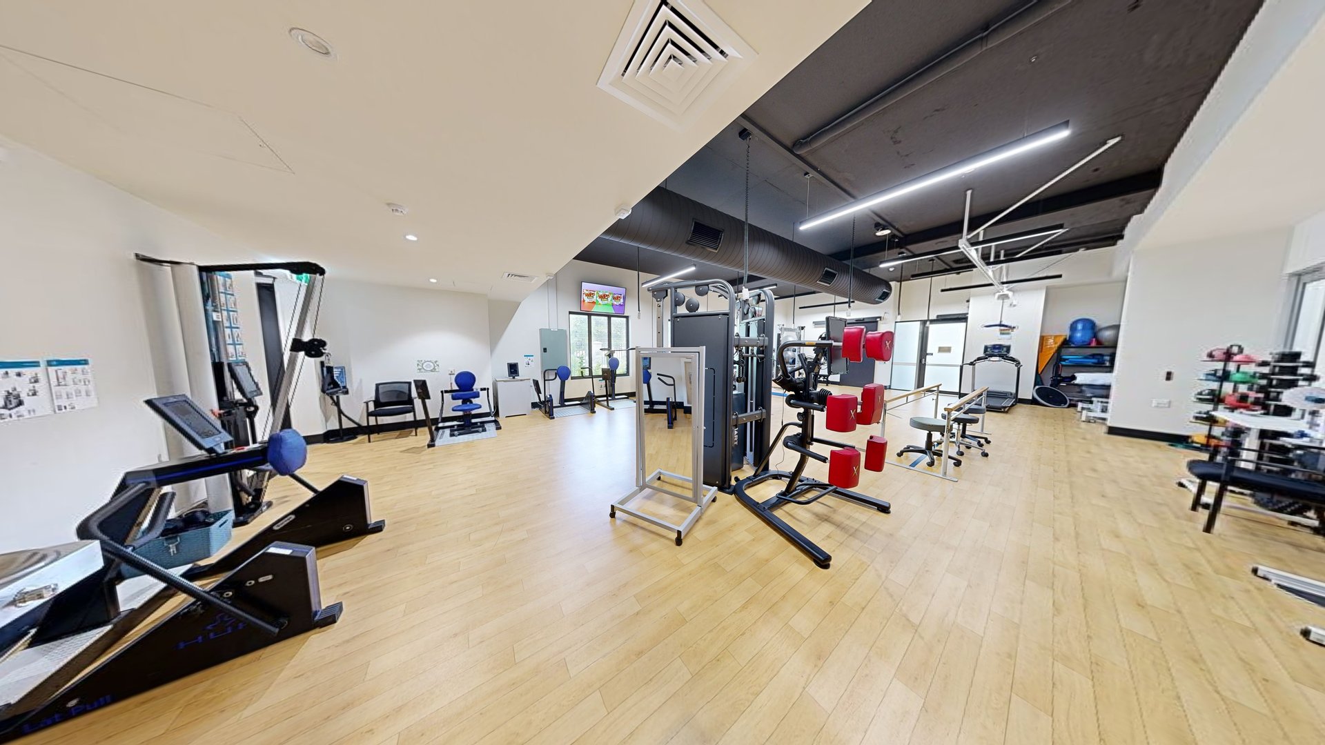 A lobby leading to the gym. Green doors on the right lead to Suites 3 and 4. On the left, an outward-opening glass door with affixed posters. A hand sanitiser machine is placed before it. Inside the gym, machines are arranged. The floor is timber, and the ceiling and walls are all white. A lobby leading to the gym. Green doors on the right lead to Suites 3 and 4. On the left, an outward-opening glass door with affixed posters. A hand sanitiser machine is placed before it. Inside the gym, machines are arranged. The floor is timber, and the ceiling and walls are all white.