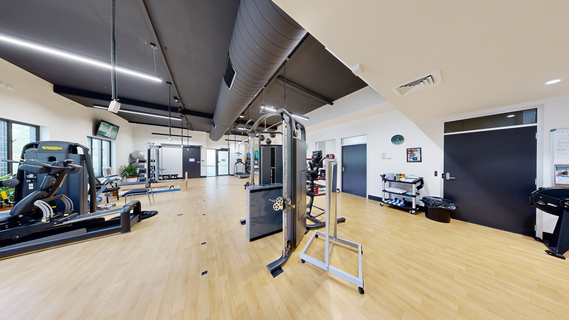 Inside the gym, a treadmill machine and bench machines are on the left, with a lat pull-down machine in the middle. The floor is timber, the walls are white with posters, and blue doors are on the right. A cart holding towels and cleaning materials sits beside a black rubbish bin against the right wall. A wall clock and poster adorn the wall above. The ceiling is black with strips of fluorescent lights and large aircon vents. Inside the gym, a treadmill machine and bench machines are on the left, with a lat pull-down machine in the middle. The floor is timber, the walls are white with posters, and blue doors are on the right. A cart holding towels and cleaning materials sits beside a black rubbish bin against the right wall. A wall clock and poster adorn the wall above. The ceiling is black with strips of fluorescent lights and large aircon vents.