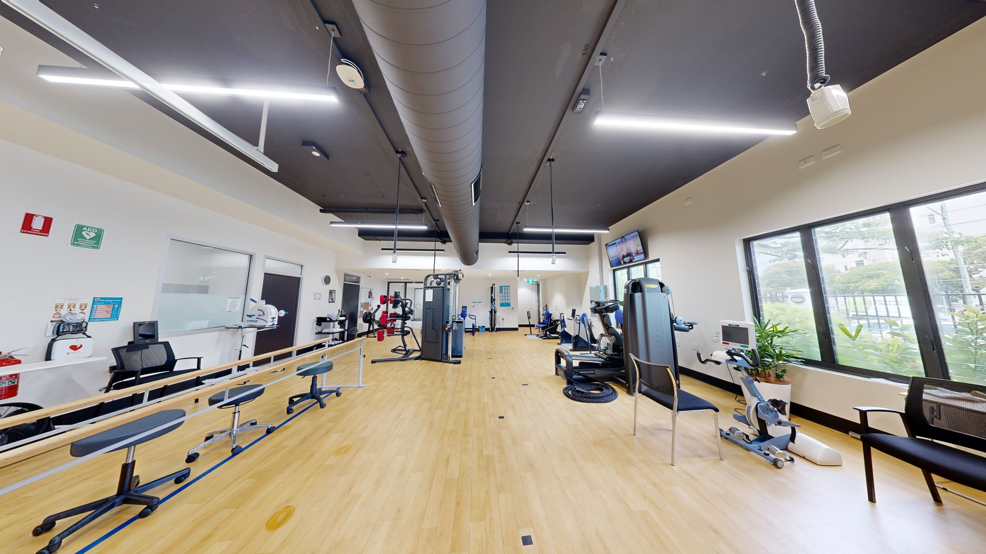 To the right is a bench chair positioned against a glass window, with Physiotherapy equipment adjacent to it. In the middle are a Smith machine and a boxing machine, with other gym equipment further in. Stools are arranged on the left side enclosed by a ribbon bollard. The walls on the left are adorned with posters, first aid equipment, and a fire extinguisher. A flatscreen TV is mounted on the upper right wall. The ceiling is black with strips of fluorescent lights and large aircon vents. To the right is a bench chair positioned against a glass window, with Physiotherapy equipment adjacent to it. In the middle are a Smith machine and a boxing machine, with other gym equipment further in. Stools are arranged on the left side enclosed by a ribbon bollard. The walls on the left are adorned with posters, first aid equipment, and a fire extinguisher. A flatscreen TV is mounted on the upper right wall. The ceiling is black with strips of fluorescent lights and large aircon vents.