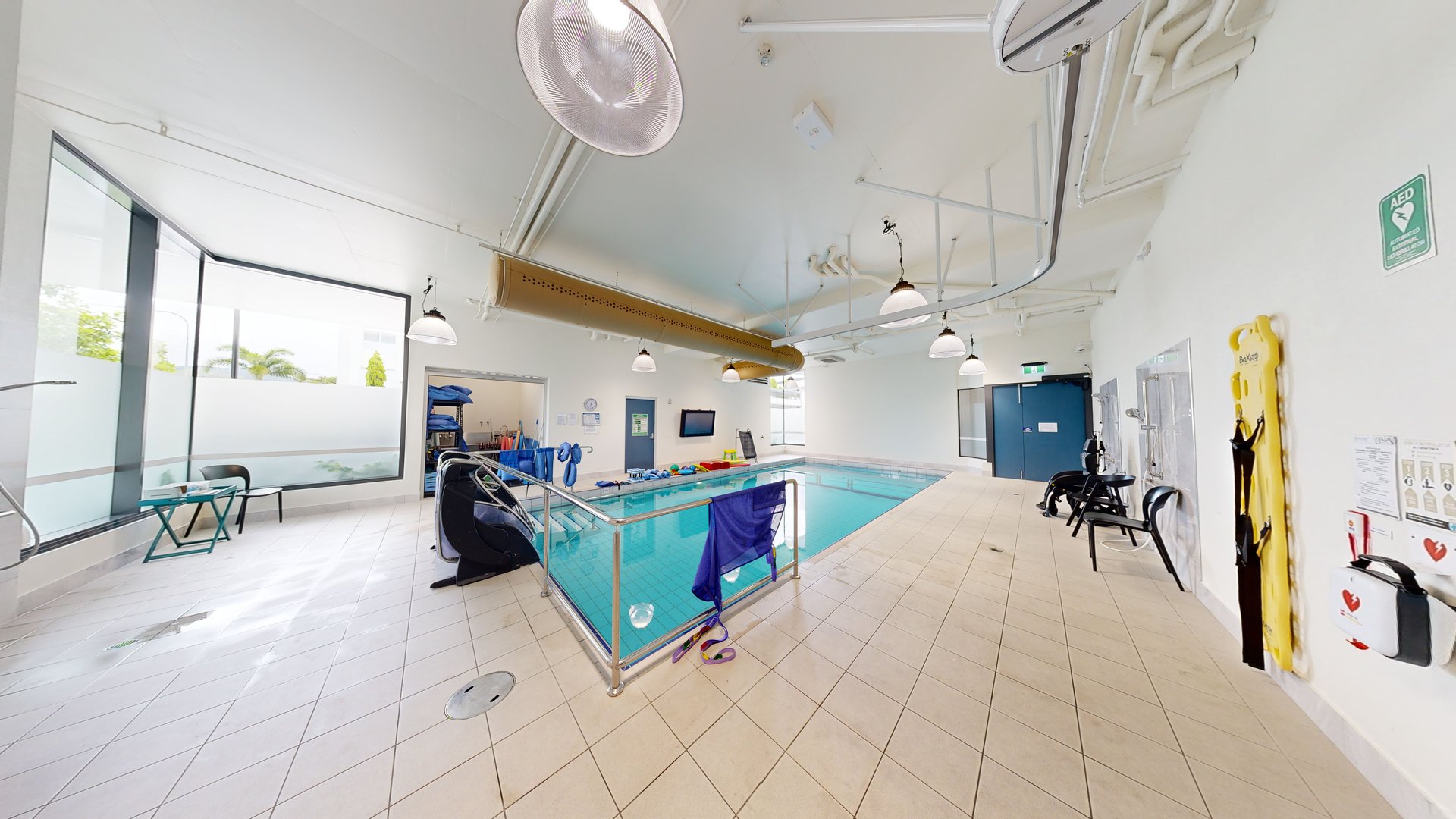 In the middle is a pool equipped with stairs and handrails. To the left is a glass wall, with chairs arranged for seating. To the right is an outdoor shower area. Aquatic wheelchairs are placed against the wall, along with posters and a first aid kit. Swimming accessories are also visible on the other end of the pool. The ceiling and walls are white with an air conditioning vent and stylish light fixtures. In the middle is a pool equipped with stairs and handrails. To the left is a glass wall, with chairs arranged for seating. To the right is an outdoor shower area. Aquatic wheelchairs are placed against the wall, along with posters and a first aid kit. Swimming accessories are also visible on the other end of the pool. The ceiling and walls are white with an air conditioning vent and stylish light fixtures.