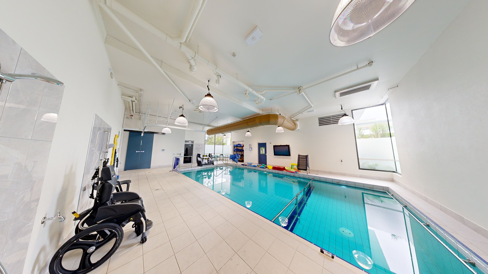In the centre is a pool with stairs and handrails. On the left is an outdoor shower area. An aquatic wheelchair is placed nearby. Swimming accessories are seen at the opposite end. The ceiling and walls are white with an air conditioning vent and stylish light fixtures. Two glass windows offer an outside view. In the centre is a pool with stairs and handrails. On the left is an outdoor shower area. An aquatic wheelchair is placed nearby. Swimming accessories are seen at the opposite end. The ceiling and walls are white with an air conditioning vent and stylish light fixtures. Two glass windows offer an outside view.