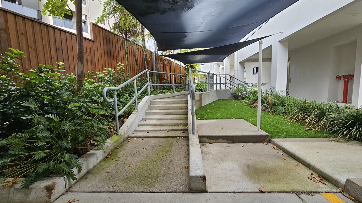 In the shelter of a corrugated roof lies accessible parking, marked by a blue wheelchair sign and yellow diagonal lines. Opposite are three vehicles of different models parked. On the right stands a white building, enclosed by a wooden fence. The area is surrounded by trees and ornamental plants. In the shelter of a corrugated roof lies accessible parking, marked by a blue wheelchair sign and yellow diagonal lines. Opposite are three vehicles of different models parked. On the right stands a white building, enclosed by a wooden fence. The area is surrounded by trees and ornamental plants.