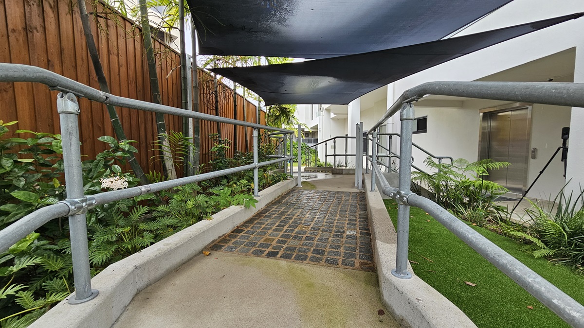 In the middle is a concrete ramp made of black tiles with metal handrails. Landscape plants and manicured grass line both sides. Overhead, a black shade cloth offers cover. The ramp descends to stairs on the right, with a lift nearby. On the left, a wooden plank fence encloses the area. In the middle is a concrete ramp made of black tiles with metal handrails. Landscape plants and manicured grass line both sides. Overhead, a black shade cloth offers cover. The ramp descends to stairs on the right, with a lift nearby. On the left, a wooden plank fence encloses the area.