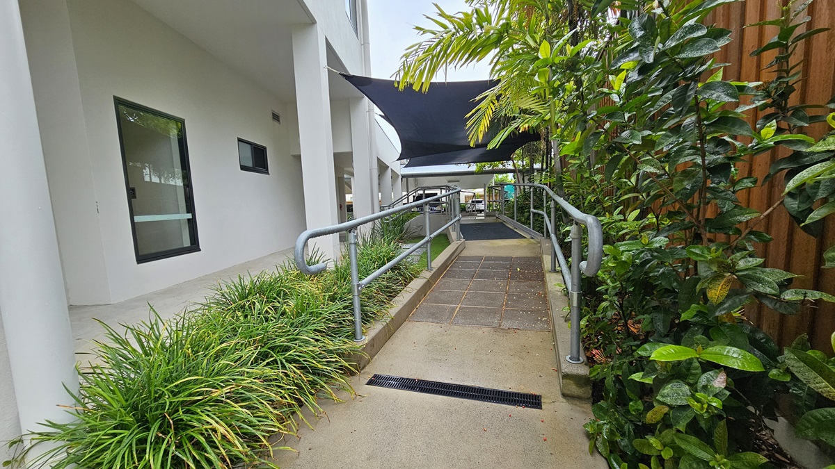 In the centre is a concrete ramp with grey tile flooring and metal handrails. Landscaped plants and manicured grass flank both sides. Above, a black shade cloth provides cover. To the right, a wooden plank fence encloses the area. On the left stands the facility building with two glass windows. In the centre is a concrete ramp with grey tile flooring and metal handrails. Landscaped plants and manicured grass flank both sides. Above, a black shade cloth provides cover. To the right, a wooden plank fence encloses the area. On the left stands the facility building with two glass windows.