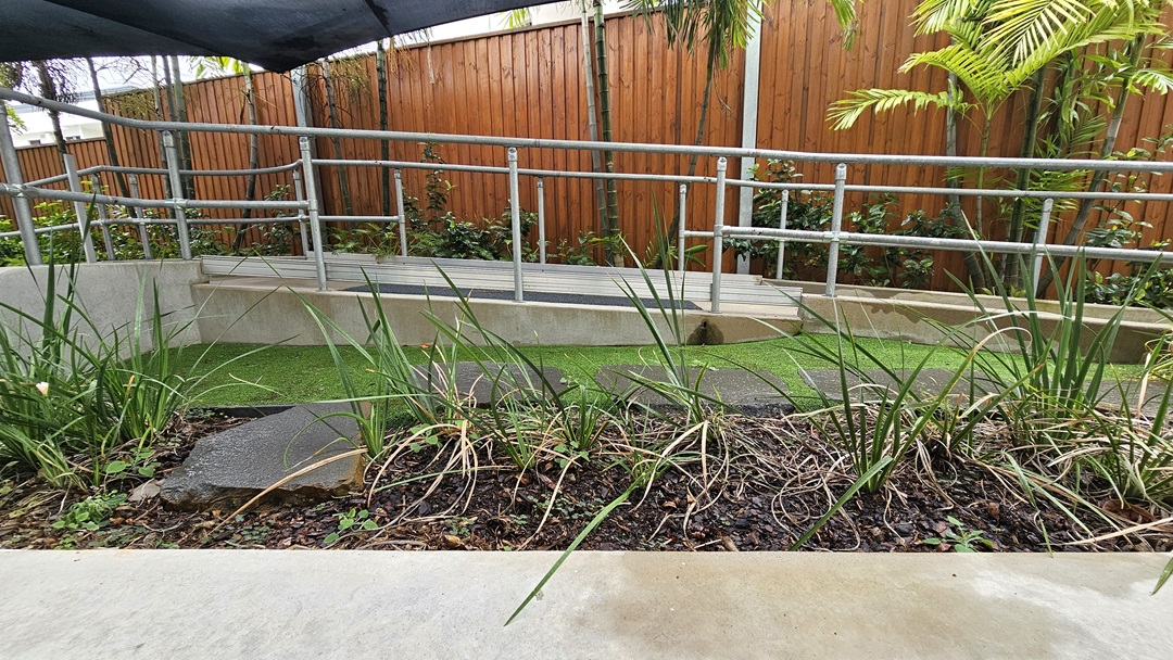 A concrete ramp with metal handrails runs between landscaped plants and manicured grass. A black shade cloth provides overhead cover, while a wooden plank fence encloses the area. A concrete ramp with metal handrails runs between landscaped plants and manicured grass. A black shade cloth provides overhead cover, while a wooden plank fence encloses the area.