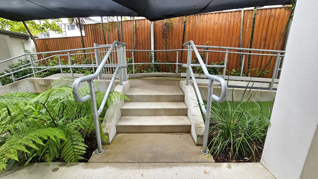 A concrete staircase with metal handrails is flanked by landscaped plants and manicured grass. Overhead, a black shade cloth provides cover, while a wooden plank fence encloses the area. A concrete staircase with metal handrails is flanked by landscaped plants and manicured grass. Overhead, a black shade cloth provides cover, while a wooden plank fence encloses the area.