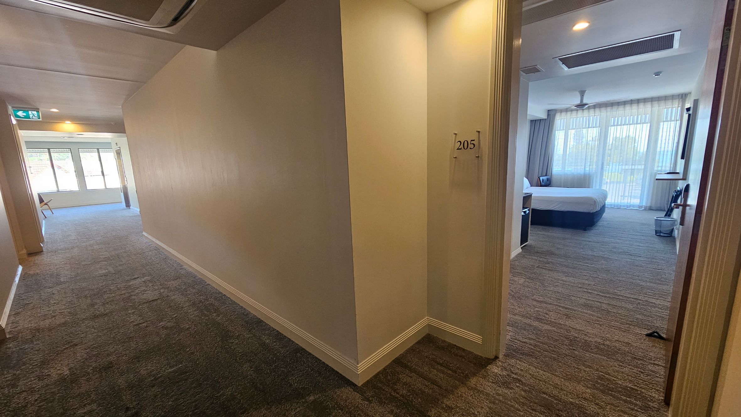 A hotel corridor with a beige carpet and cream walls. On the right, a door marked "205" is open, revealing a room with a large bed, a chair, and sheer curtains covering large windows. The ceiling has recessed lighting and an air conditioning vent. On the left, the corridor extends towards a window with a view of greenery, and an emergency exit sign is visible. A hotel corridor with a beige carpet and cream walls. On the right, a door marked "205" is open, revealing a room with a large bed, a chair, and sheer curtains covering large windows. The ceiling has recessed lighting and an air conditioning vent. On the left, the corridor extends towards a window with a view of greenery, and an emergency exit sign is visible.