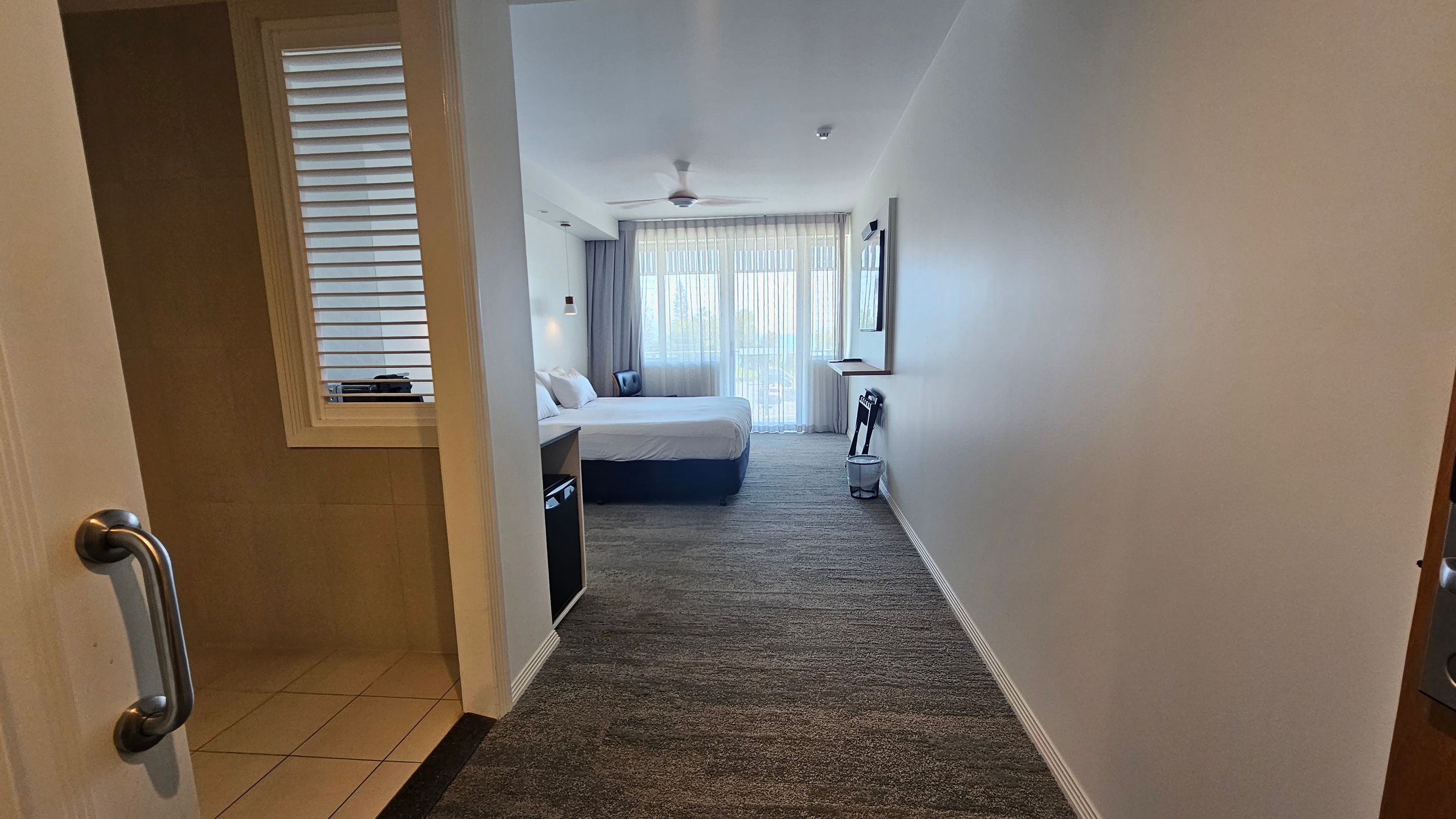 A hotel room with a view from the entrance. On the left, there's a partially open door with a metal handle leading to a tiled area. Further in, a bed with white linens is against the left wall, accompanied by a bedside table and lamp. On the right, a wall-mounted TV and a folded luggage rack are visible. The room has a carpeted floor and a ceiling fan. At the far end, sheer curtains cover a large window or balcony door, allowing natural light to enter. A hotel room with a view from the entrance. On the left, there's a partially open door with a metal handle leading to a tiled area. Further in, a bed with white linens is against the left wall, accompanied by a bedside table and lamp. On the right, a wall-mounted TV and a folded luggage rack are visible. The room has a carpeted floor and a ceiling fan. At the far end, sheer curtains cover a large window or balcony door, allowing natural light to enter.
