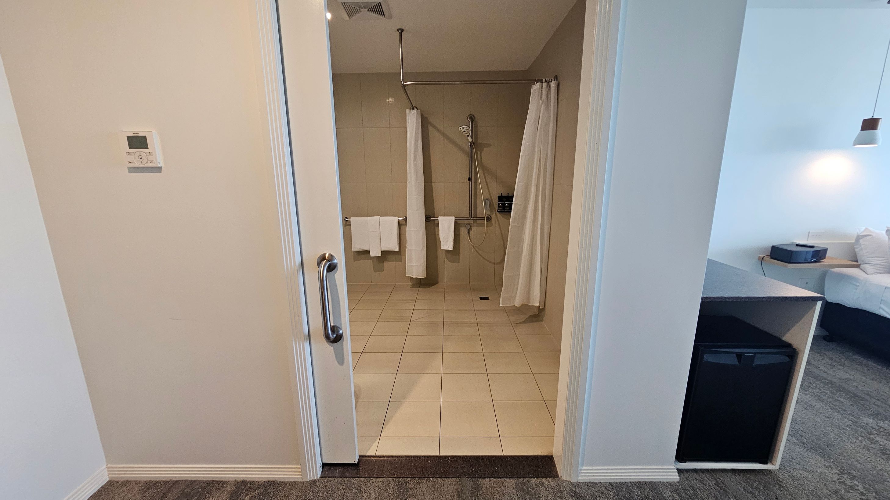 A bathroom with a tiled floor and a walk-in shower featuring a curtain and a handheld showerhead. Towels hang on a rail. On the left wall, there's a thermostat. The room outside has a carpeted floor, a small fridge under a counter, and a bed with white bedding. A pendant light hangs above the bed. A bathroom with a tiled floor and a walk-in shower featuring a curtain and a handheld showerhead. Towels hang on a rail. On the left wall, there's a thermostat. The room outside has a carpeted floor, a small fridge under a counter, and a bed with white bedding. A pendant light hangs above the bed.