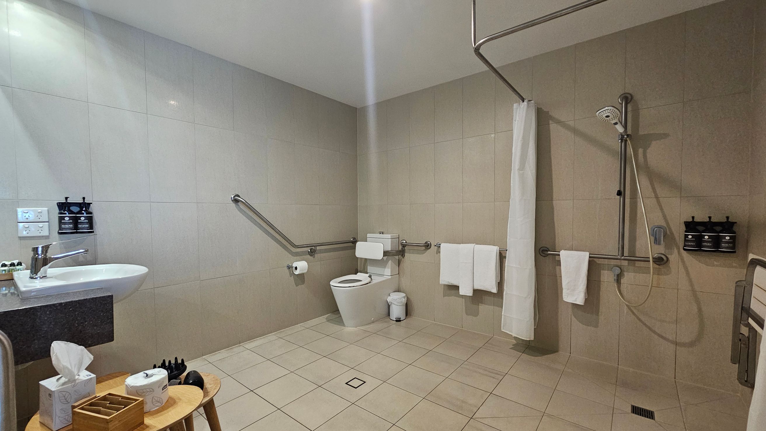 A spacious bathroom with beige tiled walls and floor. On the left, a white sink with a silver tap is mounted on a dark countertop, with soap dispensers and a light switch nearby. Below, a wooden stool holds a tissue box and toiletries. In the centre, a toilet with a support rail and a small bin. On the right, a walk-in shower with a handheld showerhead, a white curtain, and wall-mounted soap dispensers. Several grab bars and white towels are present throughout. A spacious bathroom with beige tiled walls and floor. On the left, a white sink with a silver tap is mounted on a dark countertop, with soap dispensers and a light switch nearby. Below, a wooden stool holds a tissue box and toiletries. In the centre, a toilet with a support rail and a small bin. On the right, a walk-in shower with a handheld showerhead, a white curtain, and wall-mounted soap dispensers. Several grab bars and white towels are present throughout.