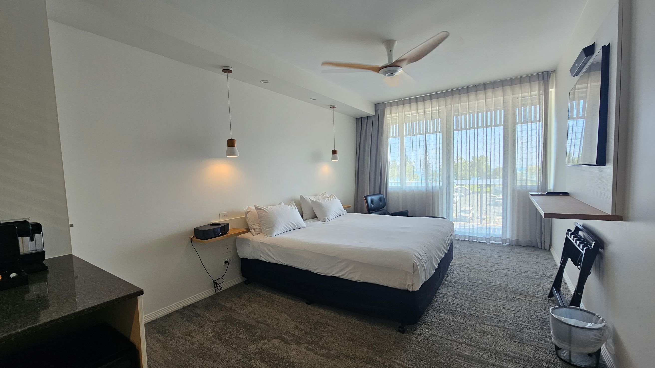 A modern hotel room with a large bed in the centre, adorned with white linens and pillows. Two pendant lights hang above the bed. On the left, a black countertop with a coffee machine. To the right, a window with sheer curtains allows natural light in, next to a blue chair. A wall-mounted TV and a small desk are on the right wall. The floor is carpeted in a mix of green and grey tones, and a ceiling fan is above. A small waste bin is on the floor near the desk. A modern hotel room with a large bed in the centre, adorned with white linens and pillows. Two pendant lights hang above the bed. On the left, a black countertop with a coffee machine. To the right, a window with sheer curtains allows natural light in, next to a blue chair. A wall-mounted TV and a small desk are on the right wall. The floor is carpeted in a mix of green and grey tones, and a ceiling fan is above. A small waste bin is on the floor near the desk.