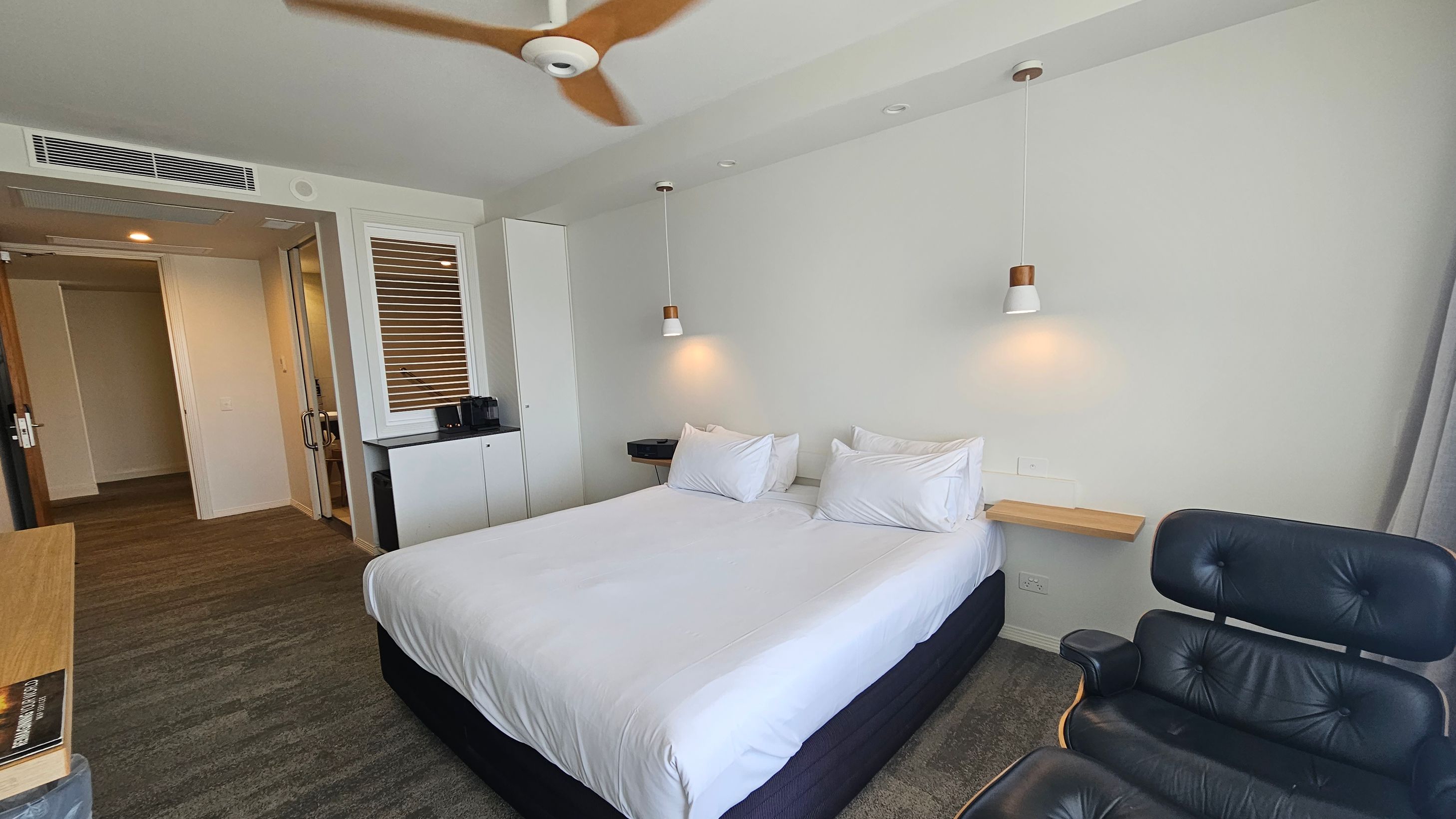 A modern hotel room with a large bed in the centre, adorned with white linens and pillows. Above the bed, two pendant lights hang from the ceiling. To the right, a black leather chair is positioned. On the left, a small cabinet with a coffee machine is placed beneath a window with blinds. The entrance to the room is visible in the background, with a wooden door and a glimpse of the bathroom. The floor is carpeted in a mix of grey and green tones, and a ceiling fan is overhead. A modern hotel room with a large bed in the centre, adorned with white linens and pillows. Above the bed, two pendant lights hang from the ceiling. To the right, a black leather chair is positioned. On the left, a small cabinet with a coffee machine is placed beneath a window with blinds. The entrance to the room is visible in the background, with a wooden door and a glimpse of the bathroom. The floor is carpeted in a mix of grey and green tones, and a ceiling fan is overhead.