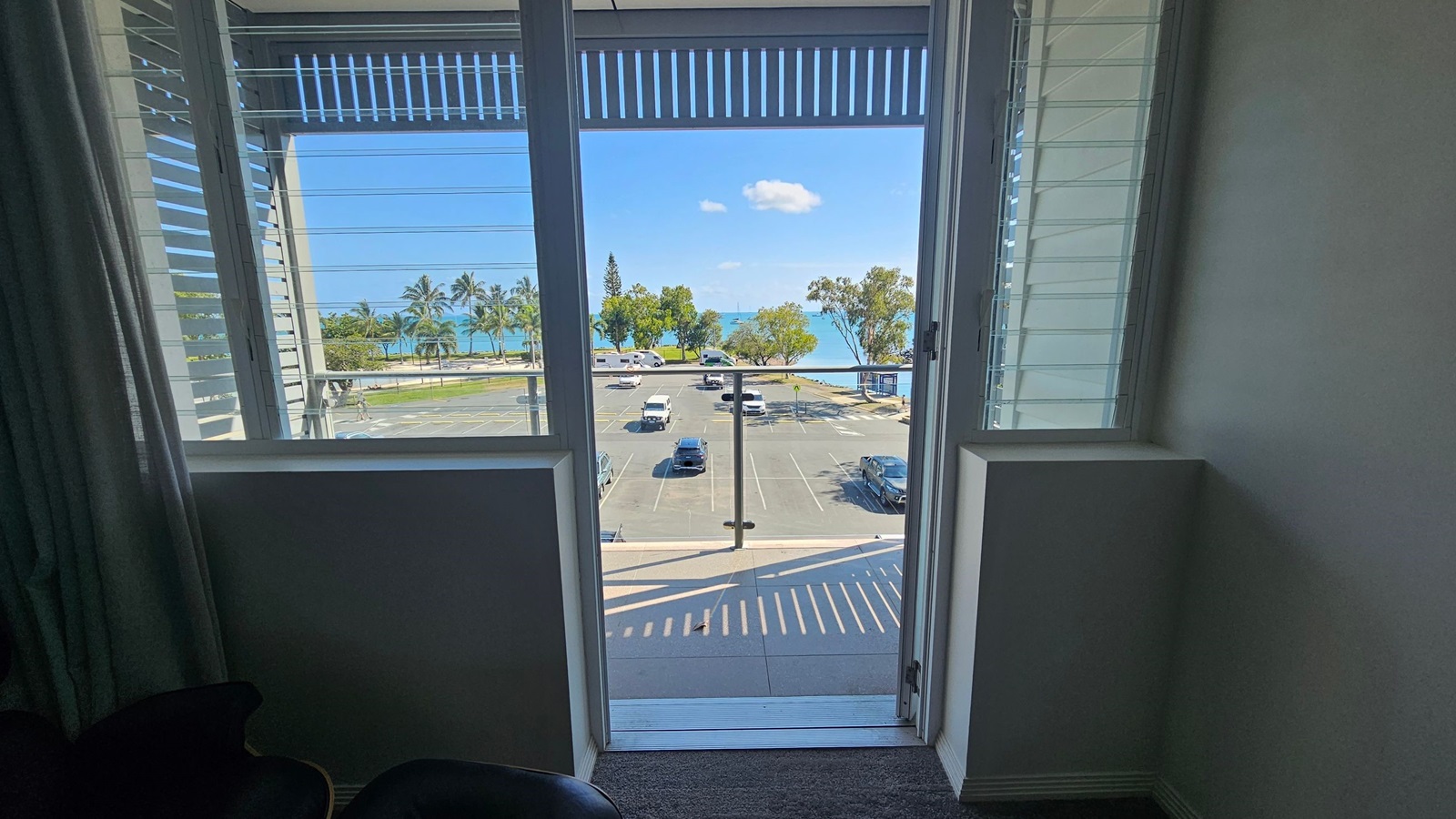 View through an open door with louvred windows on either side, leading to a balcony overlooking a car park. Beyond the car park, there are palm trees and a glimpse of the sea. The interior has a dark carpeted floor and light-coloured walls. View through an open door with louvred windows on either side, leading to a balcony overlooking a car park. Beyond the car park, there are palm trees and a glimpse of the sea. The interior has a dark carpeted floor and light-coloured walls.