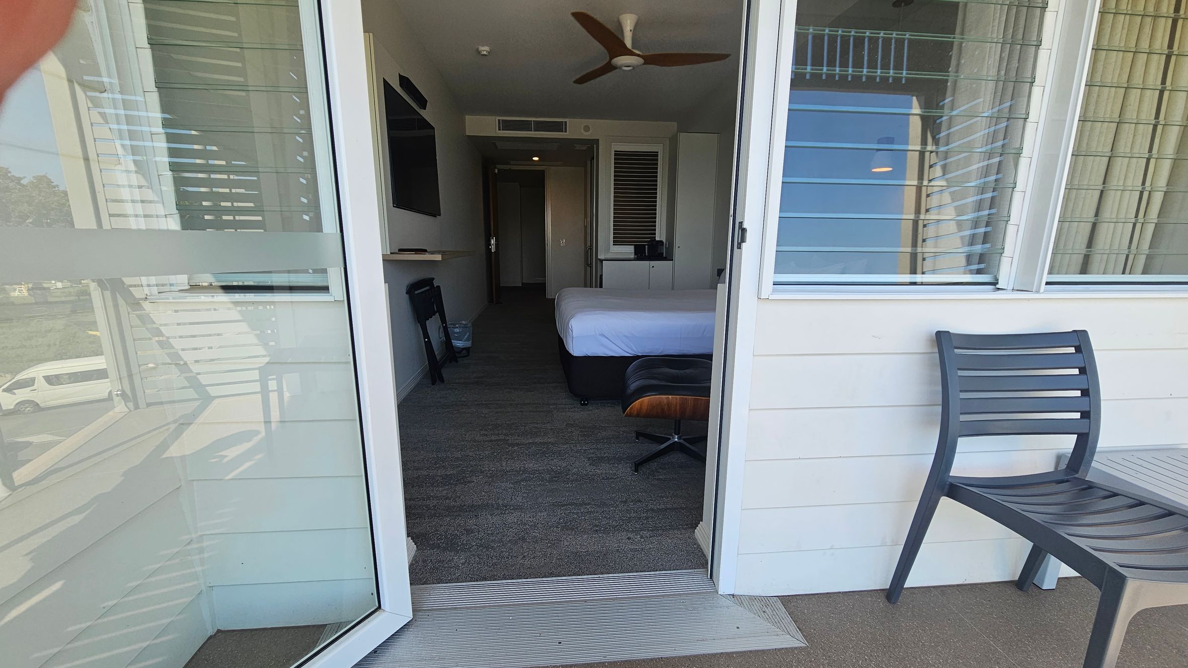 A view from a balcony into a hotel room. On the right, a dark grey chair is placed against the wall. The room has a bed with white sheets, a brown chair, and a flat-screen TV mounted on the left wall. A ceiling fan is visible above. The floor is carpeted, and there is a small table and a bin near the TV. The room has a window with horizontal blinds. Outside, a white van is parked on a grassy area. A view from a balcony into a hotel room. On the right, a dark grey chair is placed against the wall. The room has a bed with white sheets, a brown chair, and a flat-screen TV mounted on the left wall. A ceiling fan is visible above. The floor is carpeted, and there is a small table and a bin near the TV. The room has a window with horizontal blinds. Outside, a white van is parked on a grassy area.
