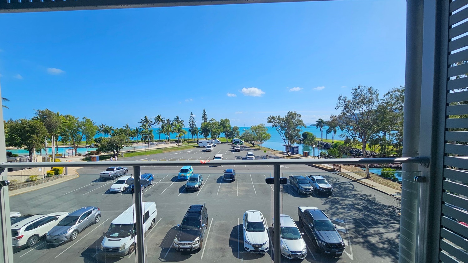View from a balcony overlooking a car park with several vehicles, including cars and a van. Beyond the car park, a road lined with palm trees leads to a beach and the sea. The sea is visible in the background, with a few small boats on the water. Trees and greenery are scattered along the shoreline. The balcony has a glass railing, and the scene is framed by vertical slats on the right. View from a balcony overlooking a car park with several vehicles, including cars and a van. Beyond the car park, a road lined with palm trees leads to a beach and the sea. The sea is visible in the background, with a few small boats on the water. Trees and greenery are scattered along the shoreline. The balcony has a glass railing, and the scene is framed by vertical slats on the right.