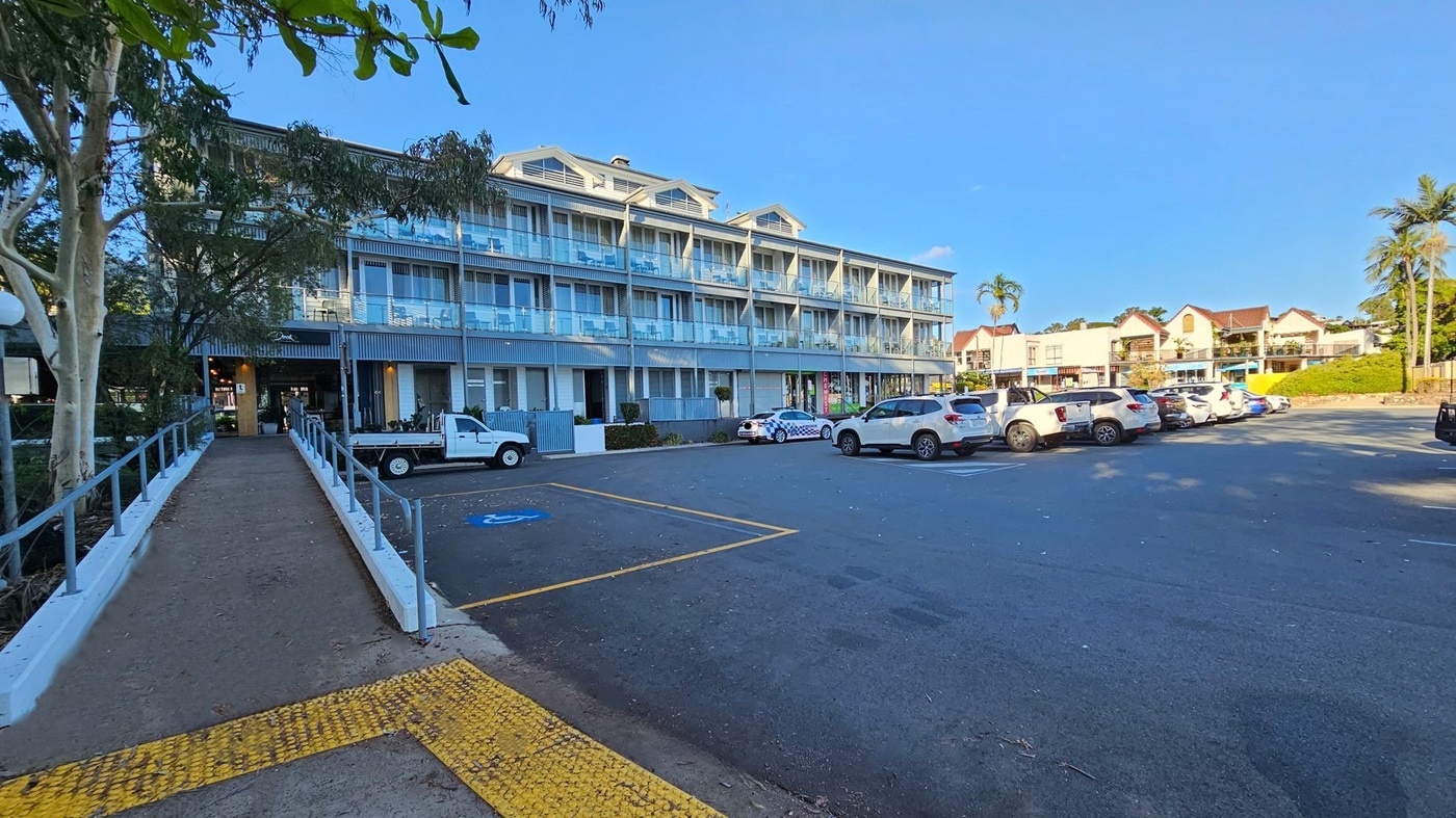 A three-storey building with a modern design, featuring glass balconies and large windows. The ground floor has a chemist shop with a visible sign. Several cars, including a police car with a checkered pattern, are parked in front. The building is surrounded by small shrubs and plants. The sky is clear and blue. A three-storey building with a modern design, featuring glass balconies and large windows. The ground floor has a chemist shop with a visible sign. Several cars, including a police car with a checkered pattern, are parked in front. The building is surrounded by small shrubs and plants. The sky is clear and blue.
