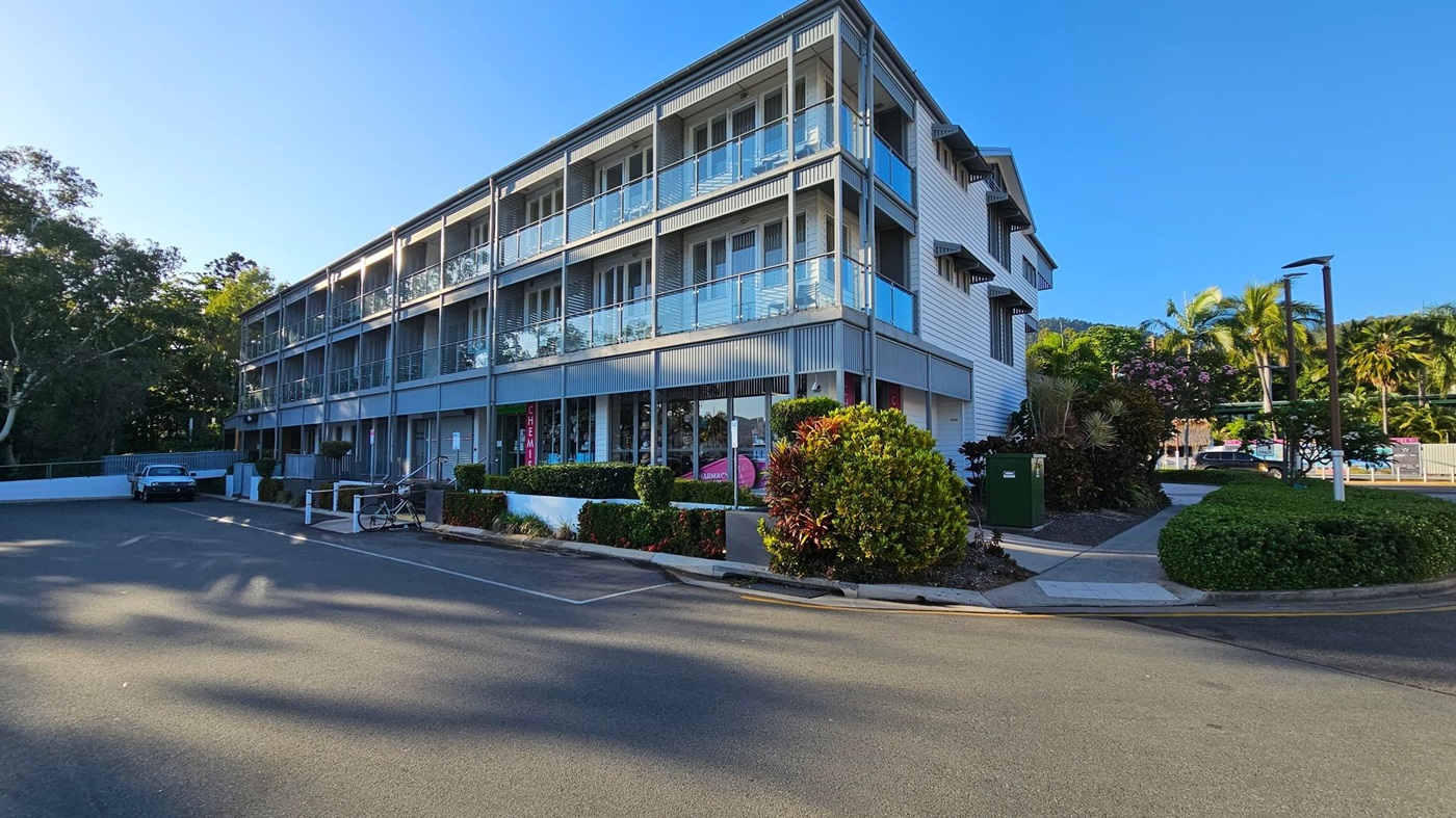 A three-storey building with a modern design featuring glass balconies and a grey facade. On the ground floor, there are shopfronts with signs, including one for a chemist. In the foreground, a paved road leads to a parking area with a parked car and a bicycle. The right side has landscaped greenery with bushes and palm trees. The background includes more trees and a clear blue sky. A three-storey building with a modern design featuring glass balconies and a grey facade. On the ground floor, there are shopfronts with signs, including one for a chemist. In the foreground, a paved road leads to a parking area with a parked car and a bicycle. The right side has landscaped greenery with bushes and palm trees. The background includes more trees and a clear blue sky.