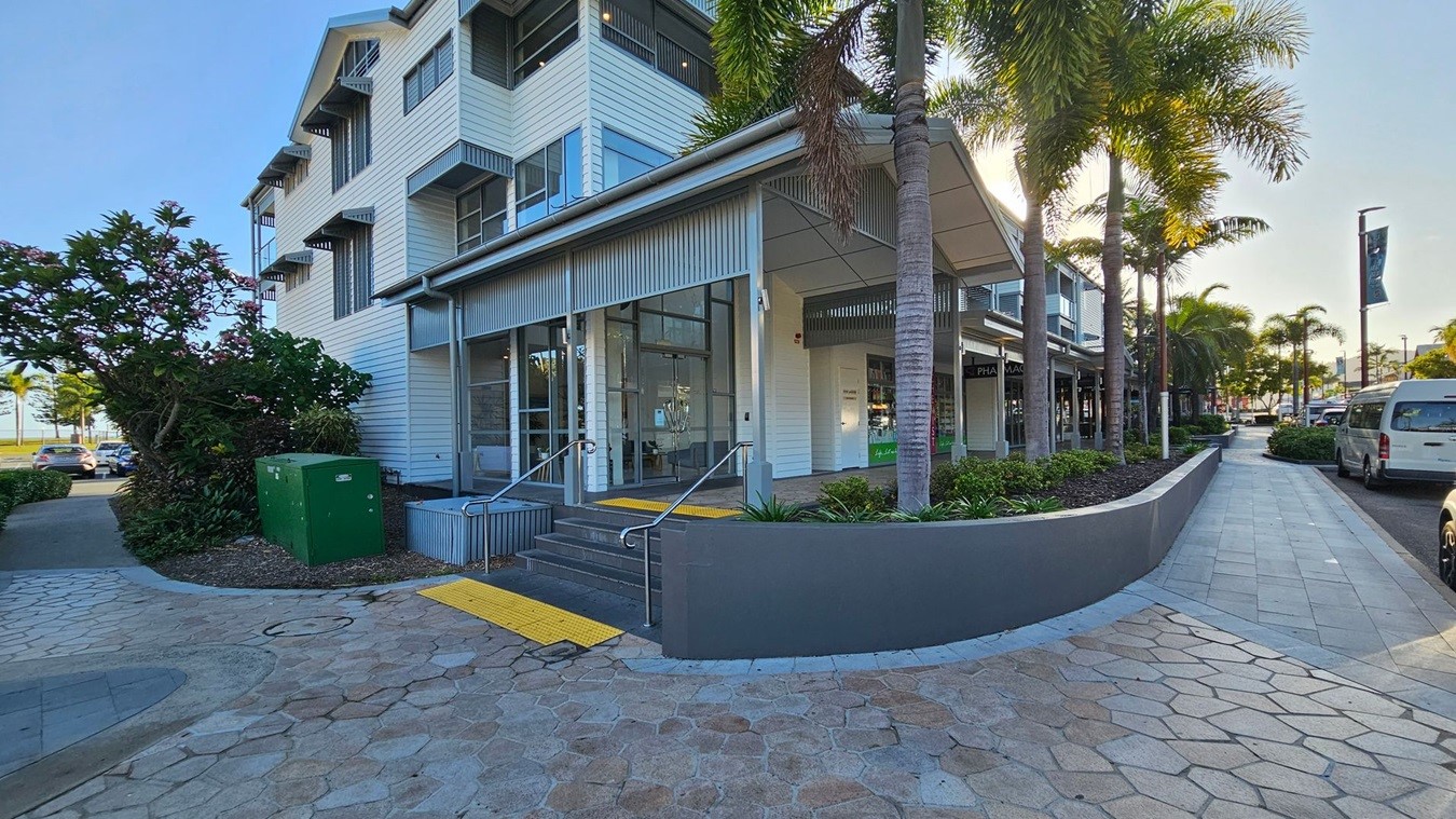 A modern building with a white exterior and large windows is on the left. It has a small set of steps leading to the entrance, with a yellow tactile paving strip. A green utility box and lush plants are nearby. On the right, a row of palm trees lines a paved walkway, with parked vehicles along the street. The pavement is made of hexagonal tiles in various shades. The building has a corrugated metal awning, and the sun is shining through the trees. A modern building with a white exterior and large windows is on the left. It has a small set of steps leading to the entrance, with a yellow tactile paving strip. A green utility box and lush plants are nearby. On the right, a row of palm trees lines a paved walkway, with parked vehicles along the street. The pavement is made of hexagonal tiles in various shades. The building has a corrugated metal awning, and the sun is shining through the trees.