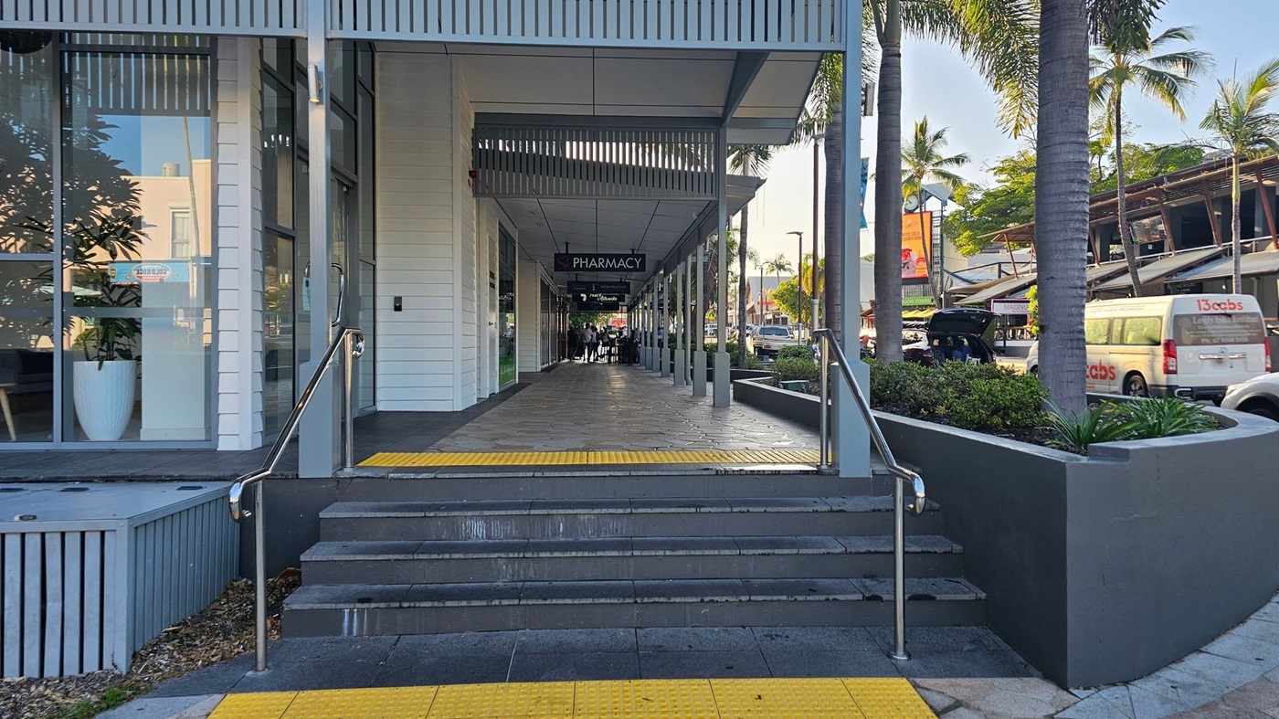 A walkway leading to a pharmacy, with steps and a ramp in the foreground. The left side features large glass windows with a potted plant inside. The right side has a raised garden bed with palm trees and a parked van. The ceiling is covered with slatted panels, and the floor is tiled. In the background, there are more shops and people walking. A walkway leading to a pharmacy, with steps and a ramp in the foreground. The left side features large glass windows with a potted plant inside. The right side has a raised garden bed with palm trees and a parked van. The ceiling is covered with slatted panels, and the floor is tiled. In the background, there are more shops and people walking.