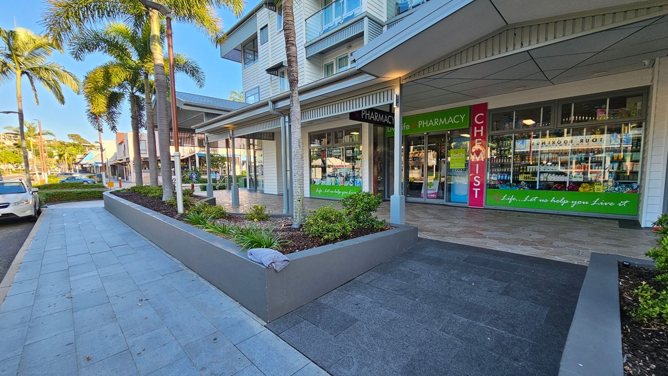 A street scene with a pharmacy on the right, featuring large glass windows displaying products. The entrance is marked by signs reading "PHARMACY" and "CHEMIST" in green and red. In the foreground, a raised garden bed with small plants and a palm tree is visible. On the left, a paved walkway runs alongside parked cars and more palm trees. The building has a modern design with a covered walkway and multiple storeys. A street scene with a pharmacy on the right, featuring large glass windows displaying products. The entrance is marked by signs reading "PHARMACY" and "CHEMIST" in green and red. In the foreground, a raised garden bed with small plants and a palm tree is visible. On the left, a paved walkway runs alongside parked cars and more palm trees. The building has a modern design with a covered walkway and multiple storeys.