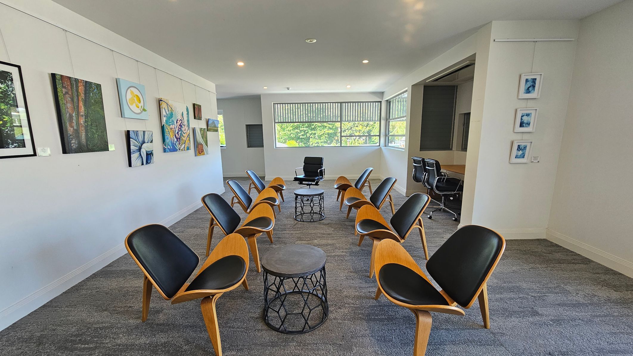 A room with a semi-circle arrangement of wooden chairs with black seats facing a single black chair and two round tables in the centre. The left wall displays various paintings, while the right wall has three small framed artworks. Large windows at the back let in natural light, and a conference table with office chairs is visible in an adjacent room. The floor is carpeted in a grey pattern, and the ceiling is plain with recessed lighting. A room with a semi-circle arrangement of wooden chairs with black seats facing a single black chair and two round tables in the centre. The left wall displays various paintings, while the right wall has three small framed artworks. Large windows at the back let in natural light, and a conference table with office chairs is visible in an adjacent room. The floor is carpeted in a grey pattern, and the ceiling is plain with recessed lighting.