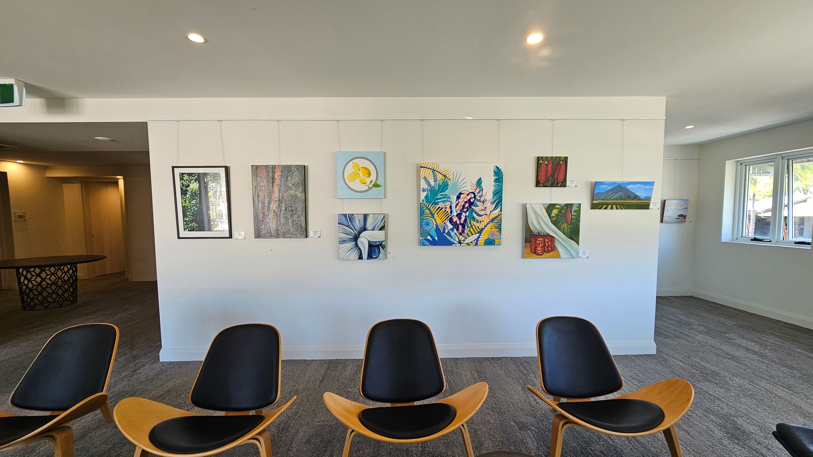 A gallery wall with various paintings, including abstract and nature-themed artworks, is displayed. Below the paintings, four modern chairs with wooden frames and black seats are arranged in a row. To the right, there are windows allowing natural light into the room. The floor is carpeted in grey, and the ceiling is white with recessed lighting. On the left, a hallway leads to another area with a round table featuring a lattice design. A gallery wall with various paintings, including abstract and nature-themed artworks, is displayed. Below the paintings, four modern chairs with wooden frames and black seats are arranged in a row. To the right, there are windows allowing natural light into the room. The floor is carpeted in grey, and the ceiling is white with recessed lighting. On the left, a hallway leads to another area with a round table featuring a lattice design.