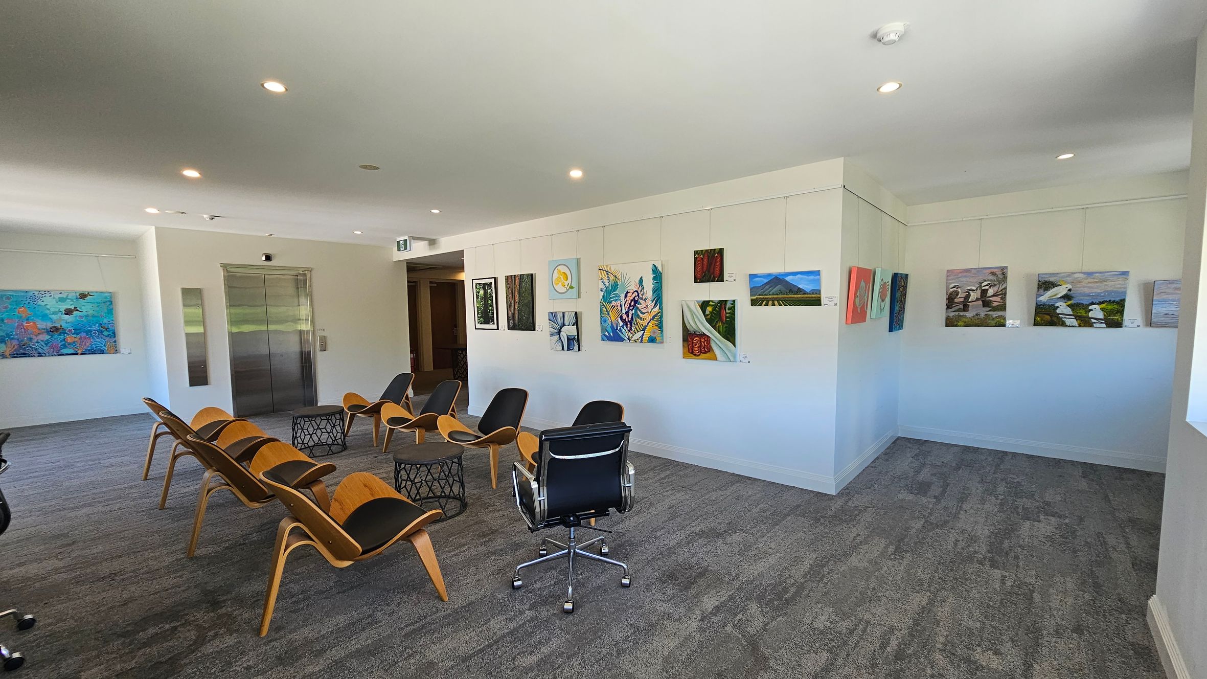 A modern gallery space with grey carpeted flooring and a white ceiling with recessed lighting. On the left, there is a wall with a colourful painting and a metal lift door. In the centre, several wooden chairs with black cushions are arranged around two small, round tables. On the right, a white wall displays various paintings, including landscapes and abstract art. A black office chair is positioned near the centre of the room. A modern gallery space with grey carpeted flooring and a white ceiling with recessed lighting. On the left, there is a wall with a colourful painting and a metal lift door. In the centre, several wooden chairs with black cushions are arranged around two small, round tables. On the right, a white wall displays various paintings, including landscapes and abstract art. A black office chair is positioned near the centre of the room.