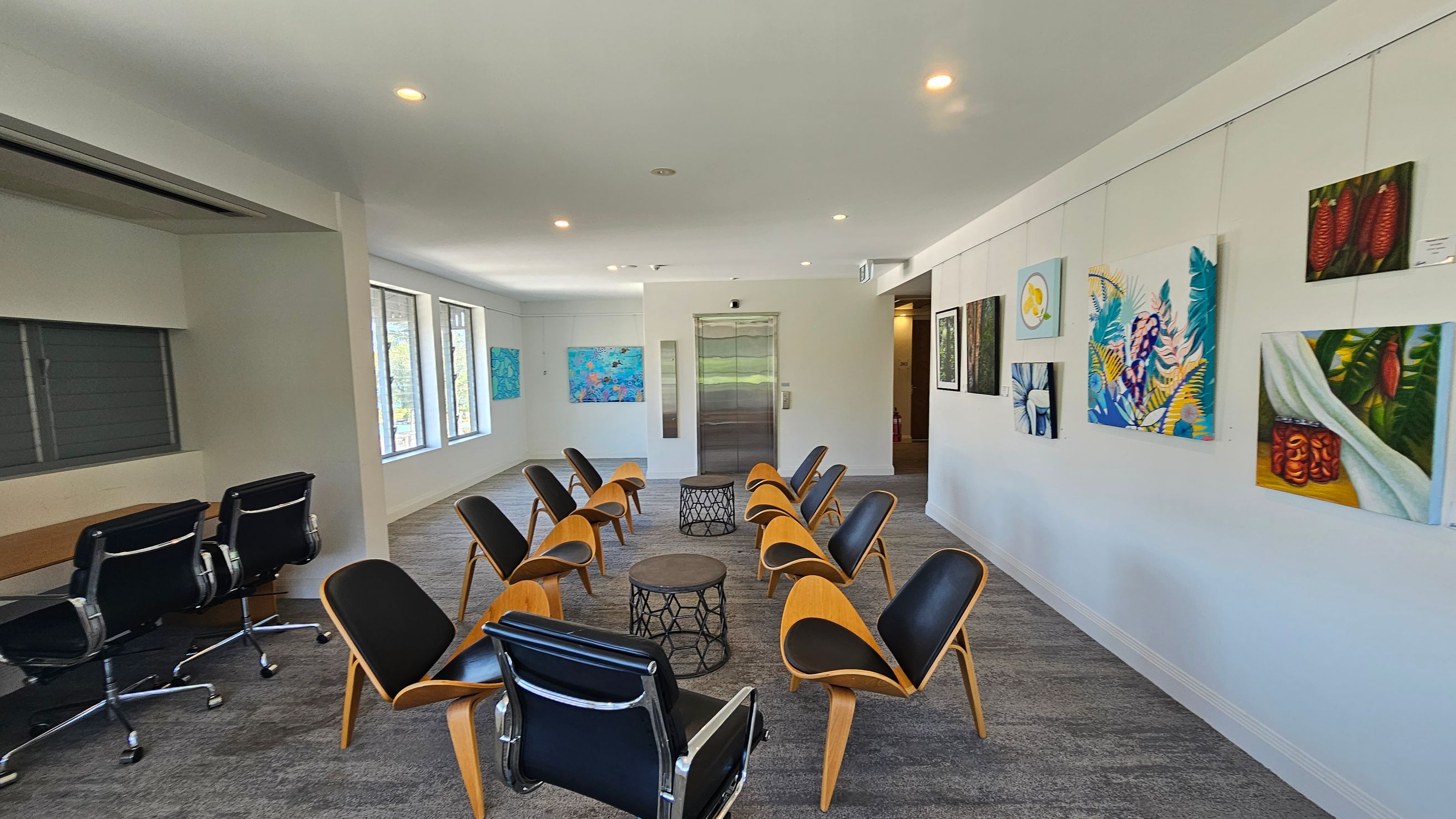 A modern room with grey carpeted flooring and a white ceiling with recessed lighting. In the centre, there are two circular tables surrounded by wooden chairs with black cushions. On the left, a row of black office chairs is positioned against a wall with windows. The right wall is adorned with colourful paintings. At the far end, an elevator is visible, flanked by more artwork. A modern room with grey carpeted flooring and a white ceiling with recessed lighting. In the centre, there are two circular tables surrounded by wooden chairs with black cushions. On the left, a row of black office chairs is positioned against a wall with windows. The right wall is adorned with colourful paintings. At the far end, an elevator is visible, flanked by more artwork.