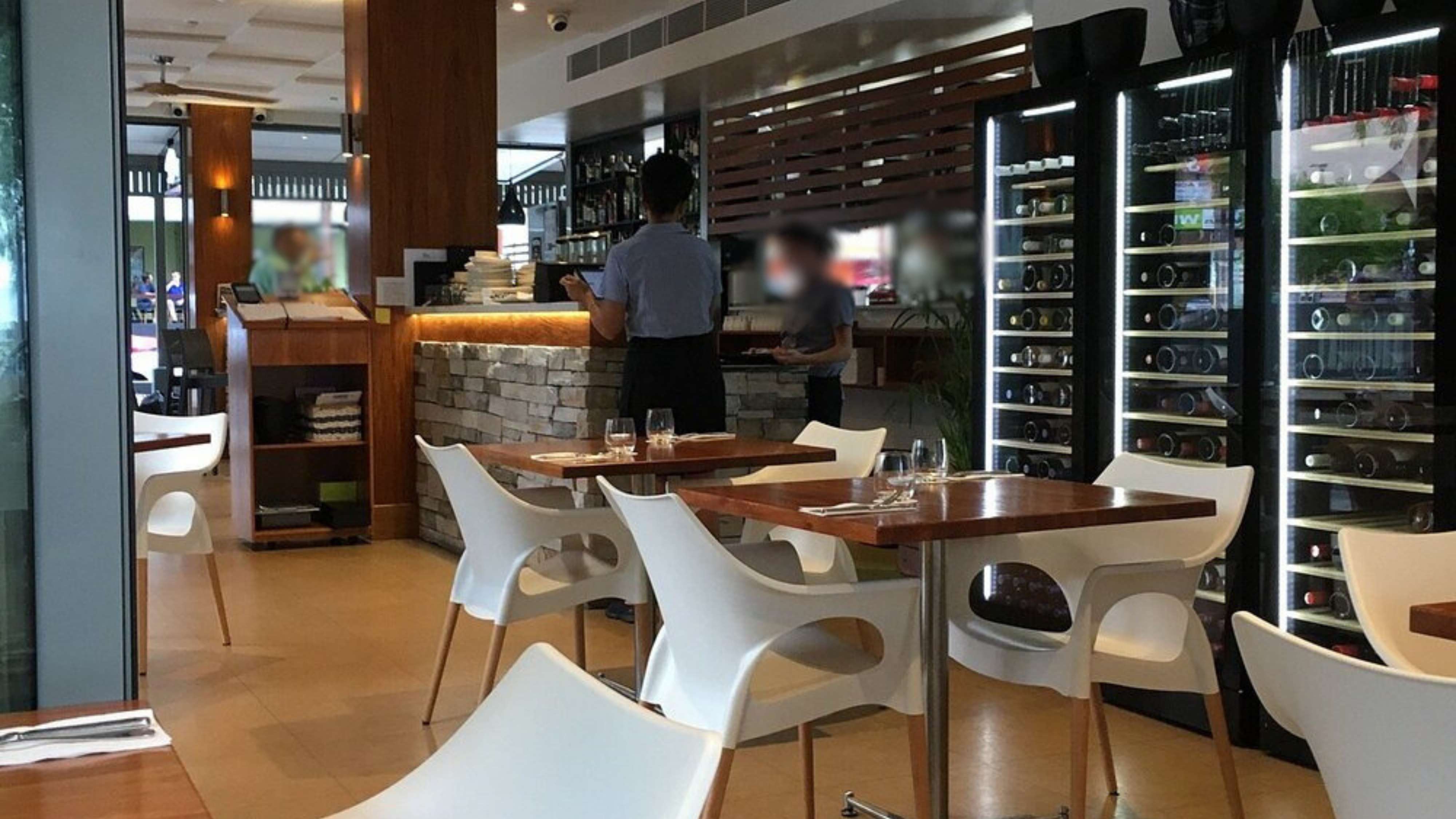 A cafe interior with a wooden panel on the right and tables in the foreground, partially visible with cutlery and glassware. To the left, there is an outdoor area with parked cars and trees. In the centre, a pathway with railings leads towards water with small boats in the distance. A red and black bicycle is parked on the right by the railings. The background features leafy trees, and the ground outside is paved with shadows present. The ceiling has slatted panels above. A cafe interior with a wooden panel on the right and tables in the foreground, partially visible with cutlery and glassware. To the left, there is an outdoor area with parked cars and trees. In the centre, a pathway with railings leads towards water with small boats in the distance. A red and black bicycle is parked on the right by the railings. The background features leafy trees, and the ground outside is paved with shadows present. The ceiling has slatted panels above.