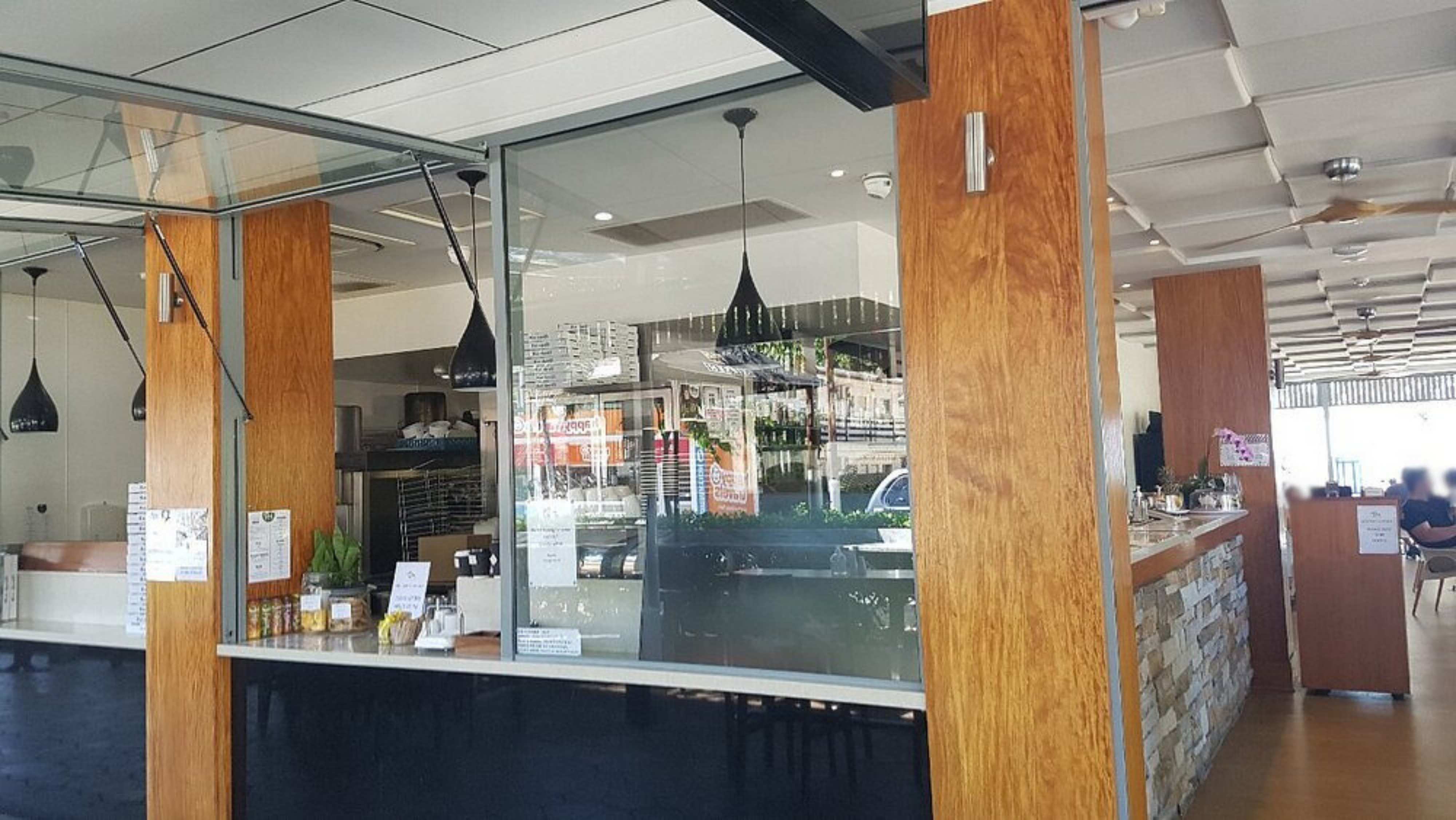 Wooden pillars are positioned on the left and right, framing glass windows. On the left, a counter displays small items, with black pendant lights hanging above. In the middle, glass panels reveal the interior, showing shelves and equipment. On the right, a stone-fronted counter runs along the wall, with tables and chairs in a dining area. The floor is light wood, and the ceiling has a grid pattern with recessed lights and ceiling fans. Wooden pillars are positioned on the left and right, framing glass windows. On the left, a counter displays small items, with black pendant lights hanging above. In the middle, glass panels reveal the interior, showing shelves and equipment. On the right, a stone-fronted counter runs along the wall, with tables and chairs in a dining area. The floor is light wood, and the ceiling has a grid pattern with recessed lights and ceiling fans.